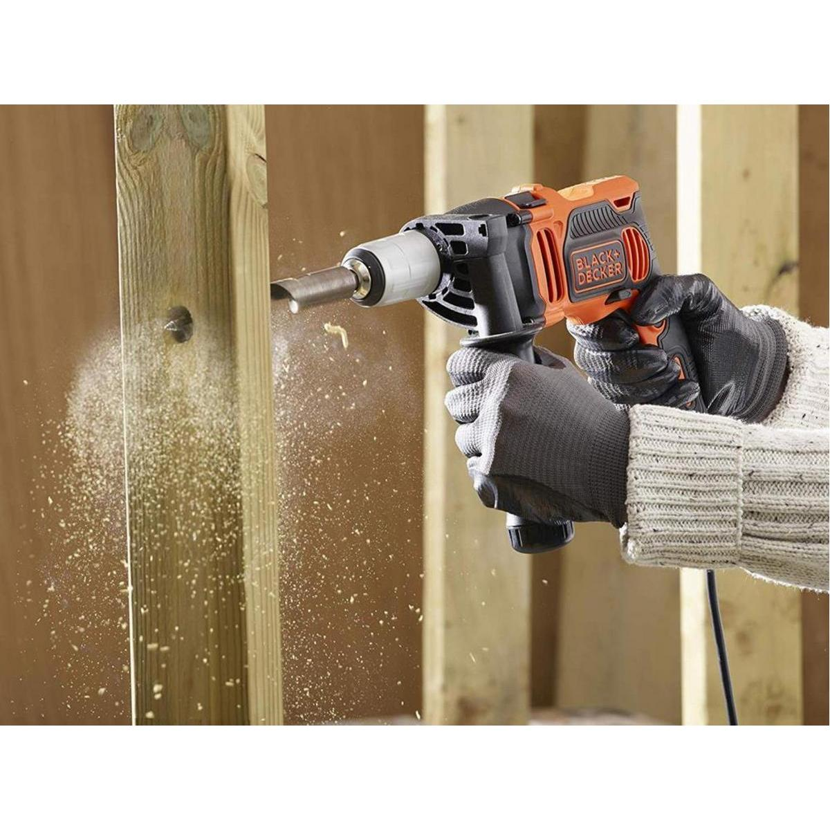 Una persona taladra madera con un taladro Black+Decker. Aserrín vuela. El taladro es naranja y negro. La persona usa guantes.