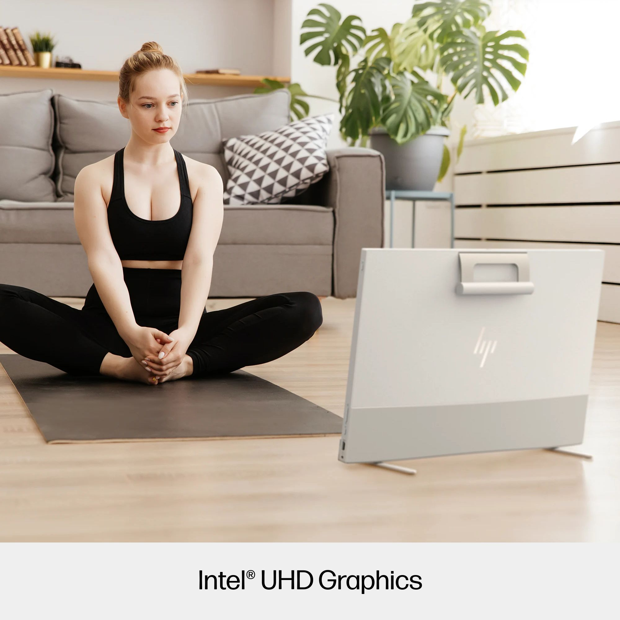 Kobieta w czarnym staniku sportowym i legginsach medytuje na macie, komputer przed nią, a w tle sofa.