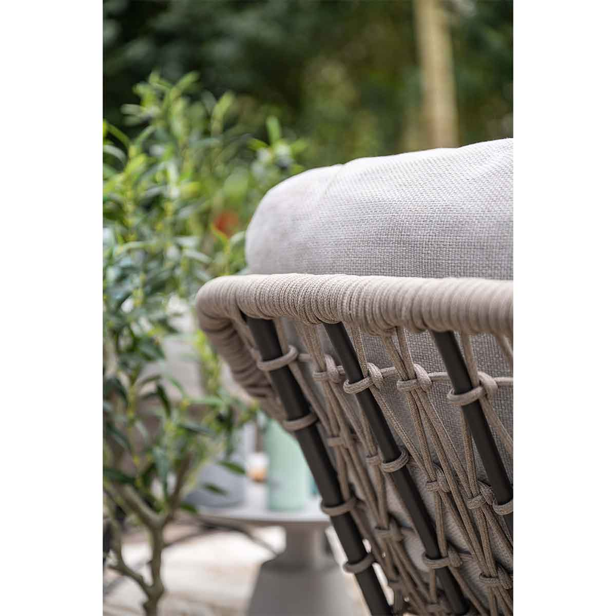 Close-up van een stoel met een geweven rugleuning, grijs kussen en touwdetails. Groen gebladerte en een tafel op de wazige achtergrond.