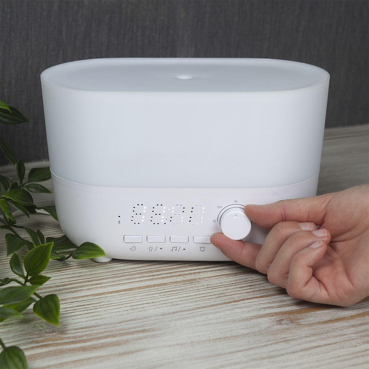 Biały dyfuzor do aromaterapii stoi na drewnianym stole. Dłoń reguluje pokrętło. Zielone liście po lewej. Tło jest szare.