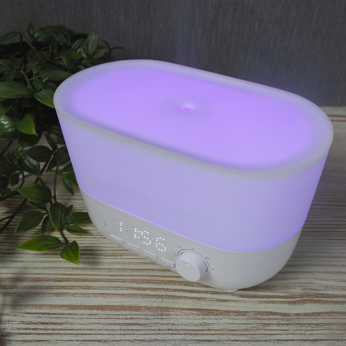 Biało-fioletowy dyfuzor do aromaterapii z cyfrowym wyświetlaczem i pokrętłem timera, na drewnianej powierzchni, z zielenią.