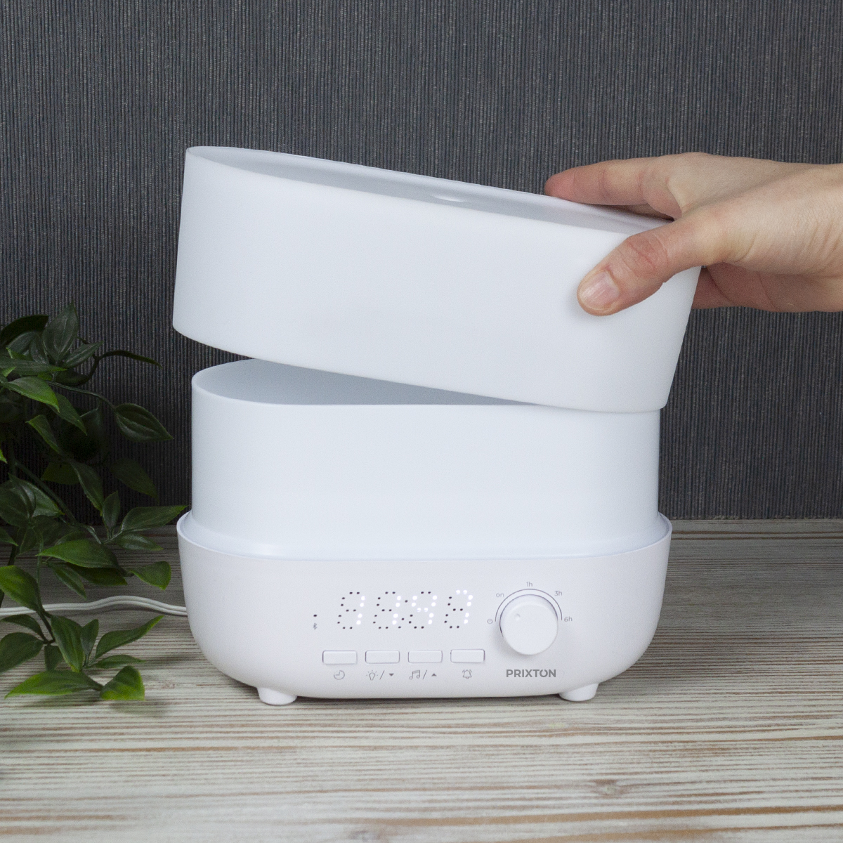 Biały nawilżacz powietrza z pokrywą podnoszoną przez dłoń. Ma wyświetlacz cyfrowy i przyciski. Zielona roślina i drewniany stół.