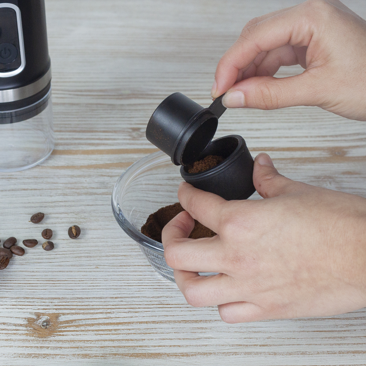 Ręce wlewają zmieloną kawę z czarnej miarki do szklanej miski. Ziarna kawy i młynek są w kadrze.