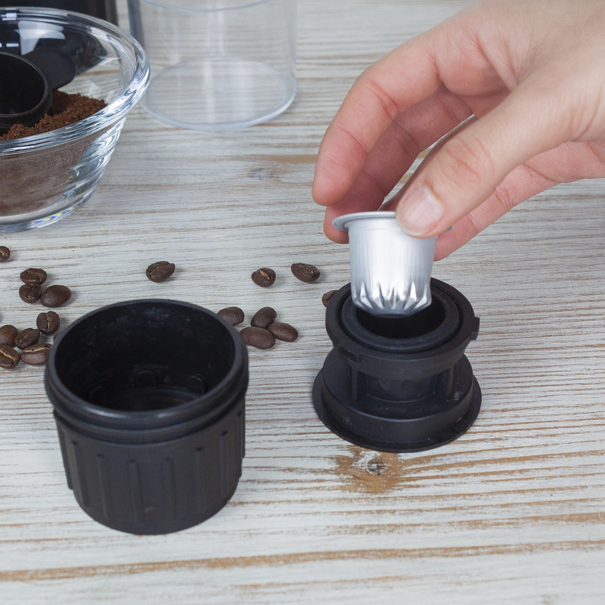 Ręka umieszcza kapsułkę z kawą w czarnym urządzeniu. Widoczne ziarna i fusy kawy. Jasne drewniane tło.