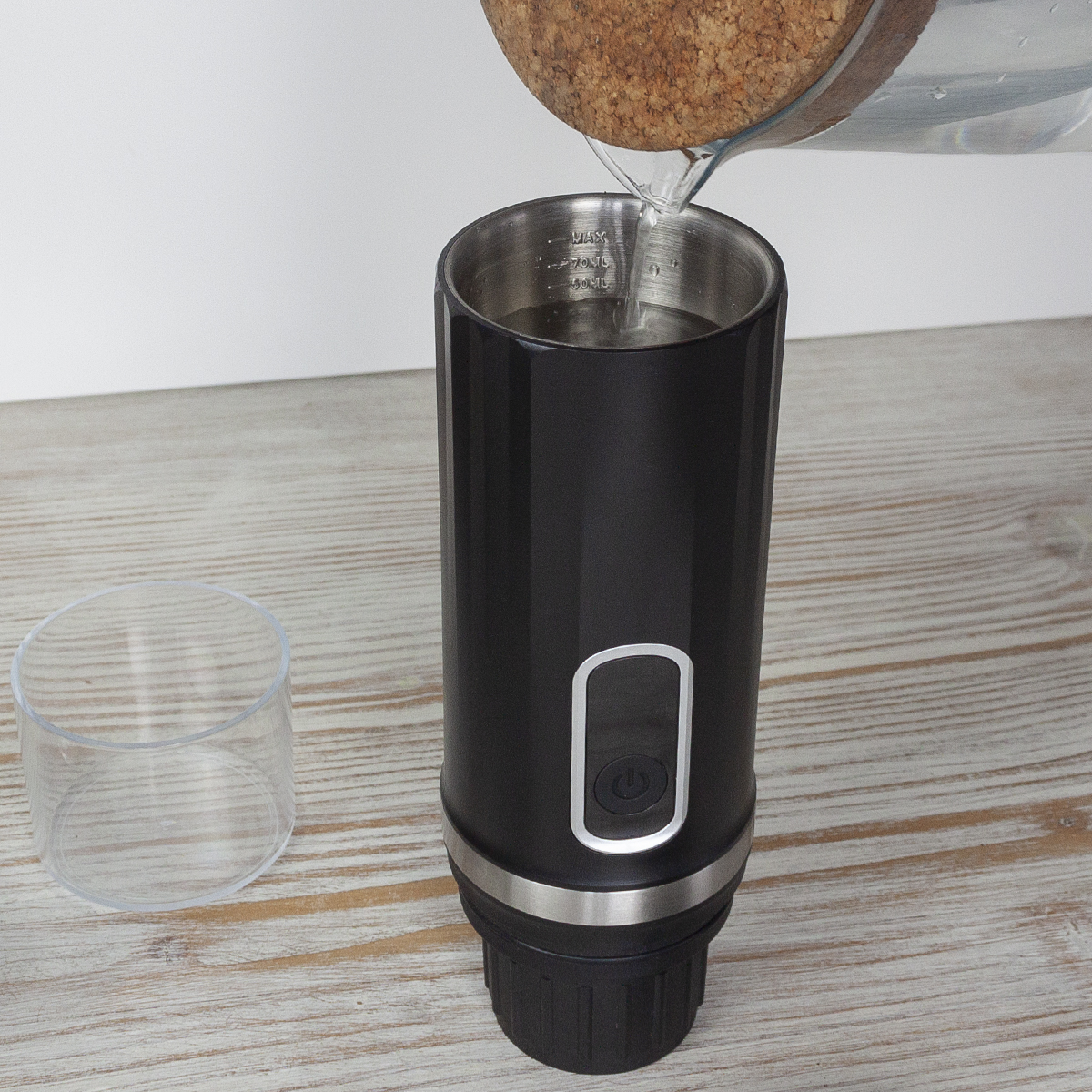Woda jest wlewana do czarnego kubka termicznego. Mały szklany kubek po lewej stronie. Tło to jasna drewniana powierzchnia i biała ściana.