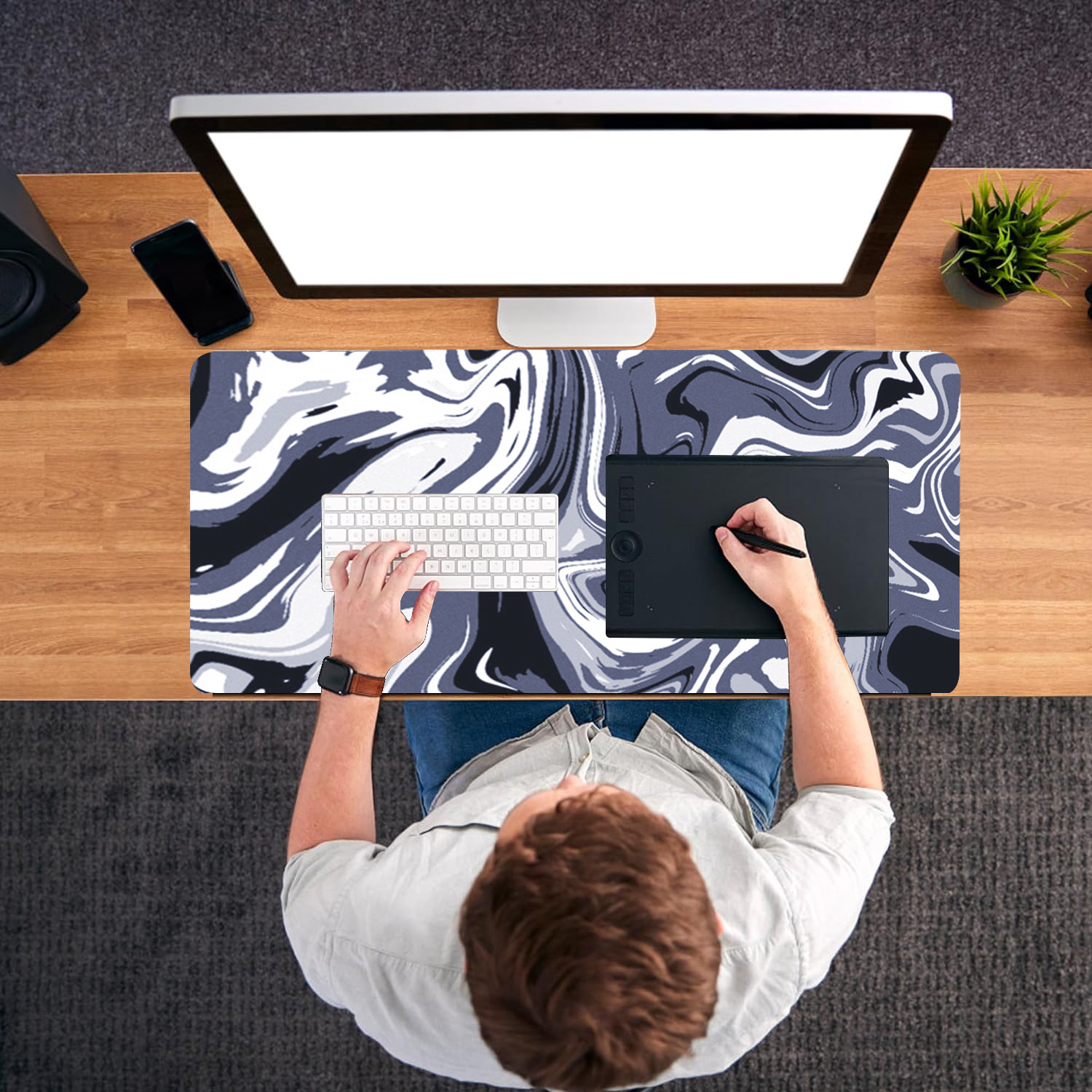 Osoba siedzi przy biurku, używając komputera z tabletem graficznym i klawiaturą. Biurko ma wzorzystą matę. Po prawej stronie znajduje się roślina.