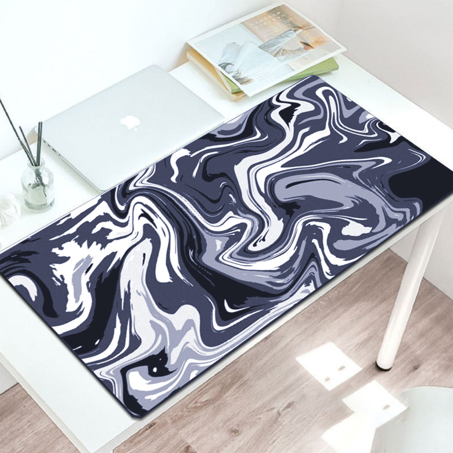 Biurko z laptopem, książkami i podkładką pod mysz z marmurowym wzorem w odcieniach szarości, bieli i czerni. Szklanka z patyczkami.