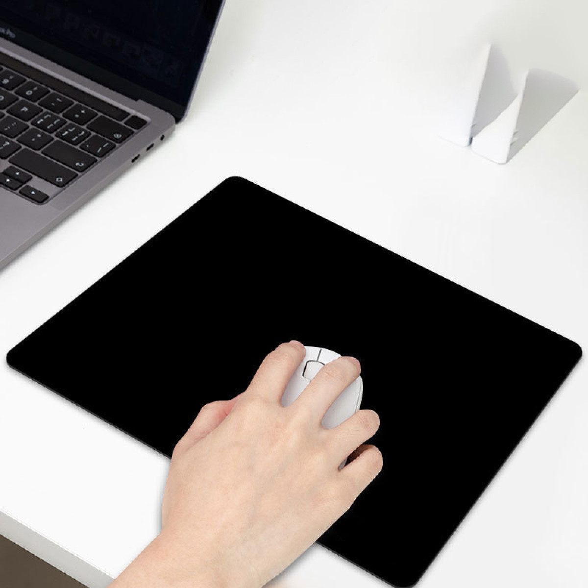 Dłoń trzyma białą mysz na czarnej podkładce. Laptop i biały przedmiot w tle na białym stole.