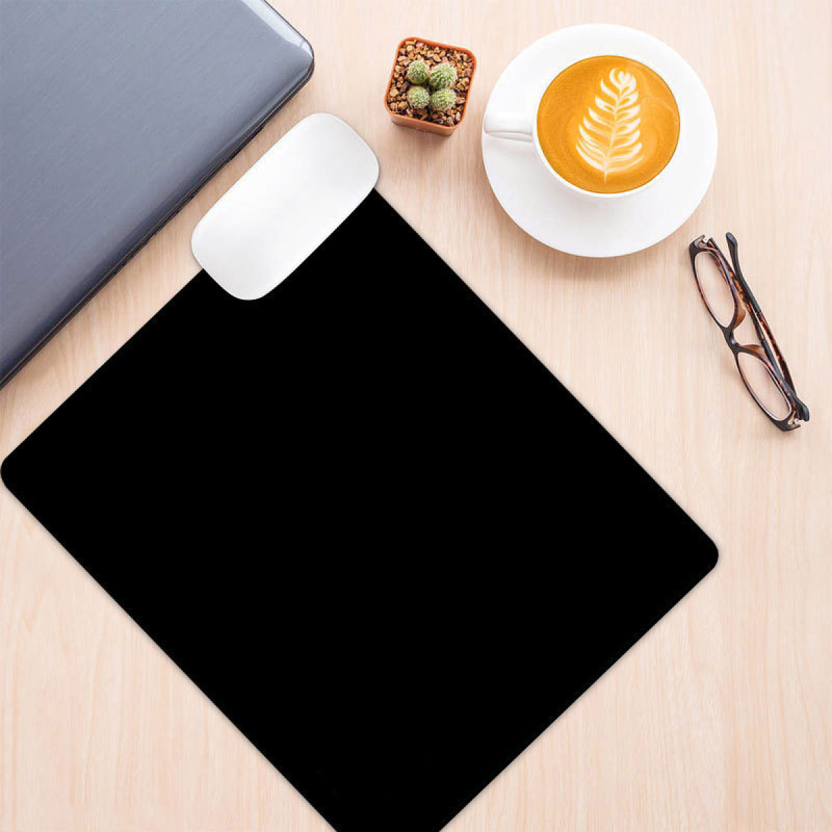 Widok z góry na biurko. Ma laptopa, mysz, czarną podkładkę pod mysz, kawę, okulary i małego kaktusa.