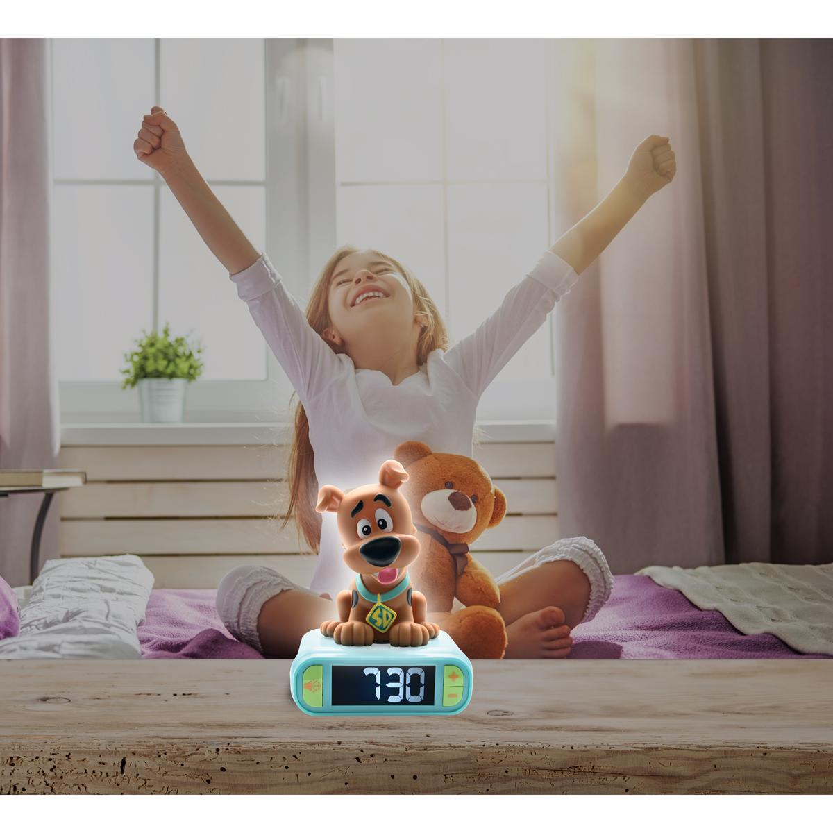 Ein Mädchen dehnt sich nach dem Aufwachen. Ein Scooby Doo Wecker und ein Teddybär liegen auf dem Bett. Die Uhrzeit ist 7:30.
