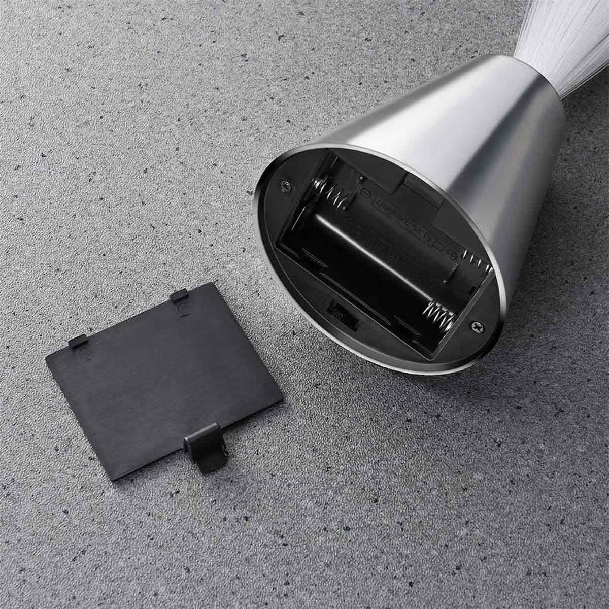 Ein silberner, kegelförmiger Lampensockel mit geöffnetem Batteriefach, abgenommener schwarzer Abdeckung und weißen Borsten auf grauem Untergrund.