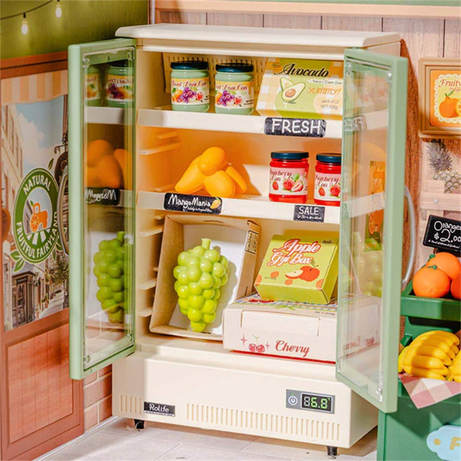 Ein Miniaturkühlschrank ist geöffnet und zeigt Obst, Gläser und Schachteln mit der Aufschrift 'Avocado', 'Mango Mania' und 'Apple Gift Box'.