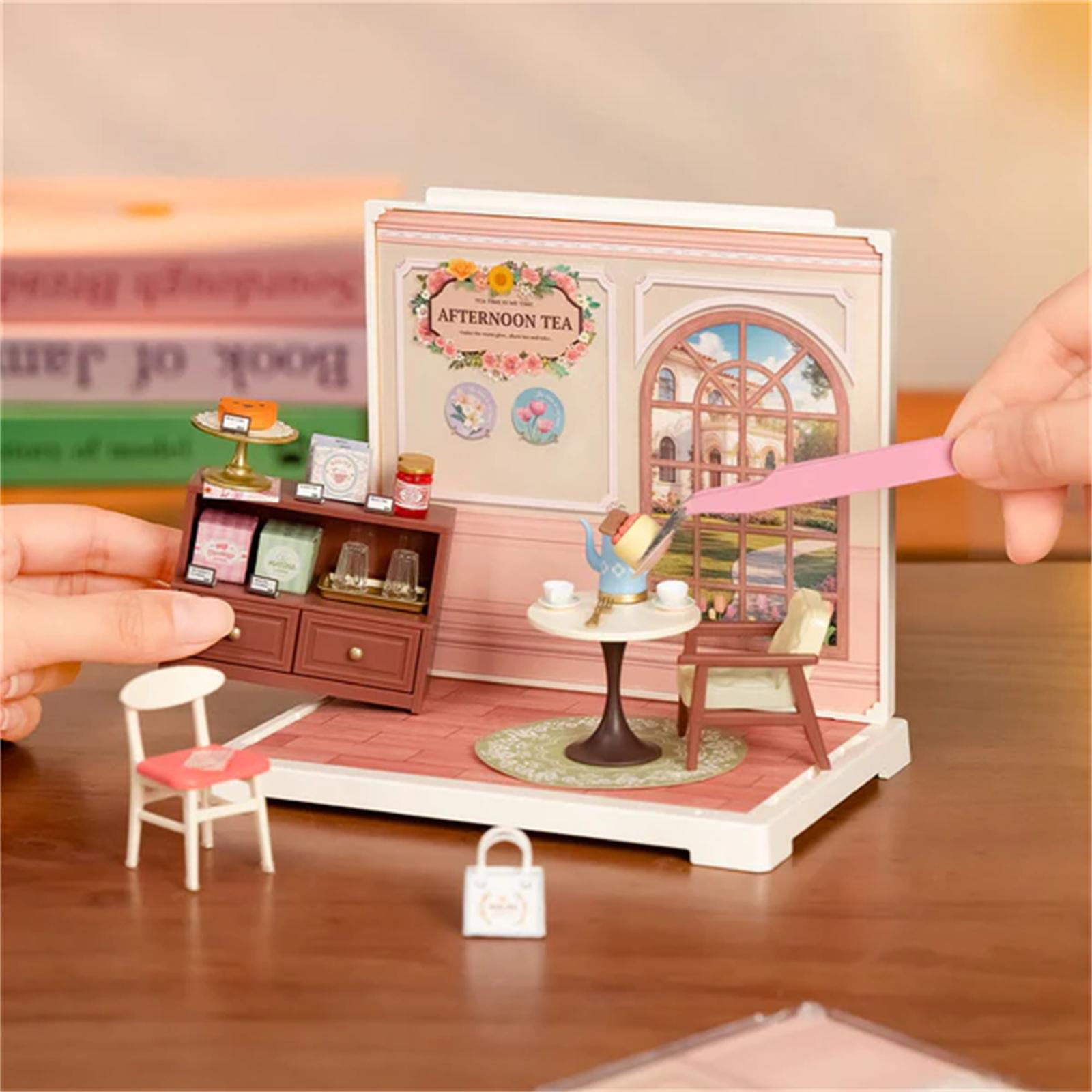 Miniatur-Nachmittagstee-Szene mit Tisch, Stühlen und Schrank. Eine Hand hält ein rosa Werkzeug in der Nähe der Szene.