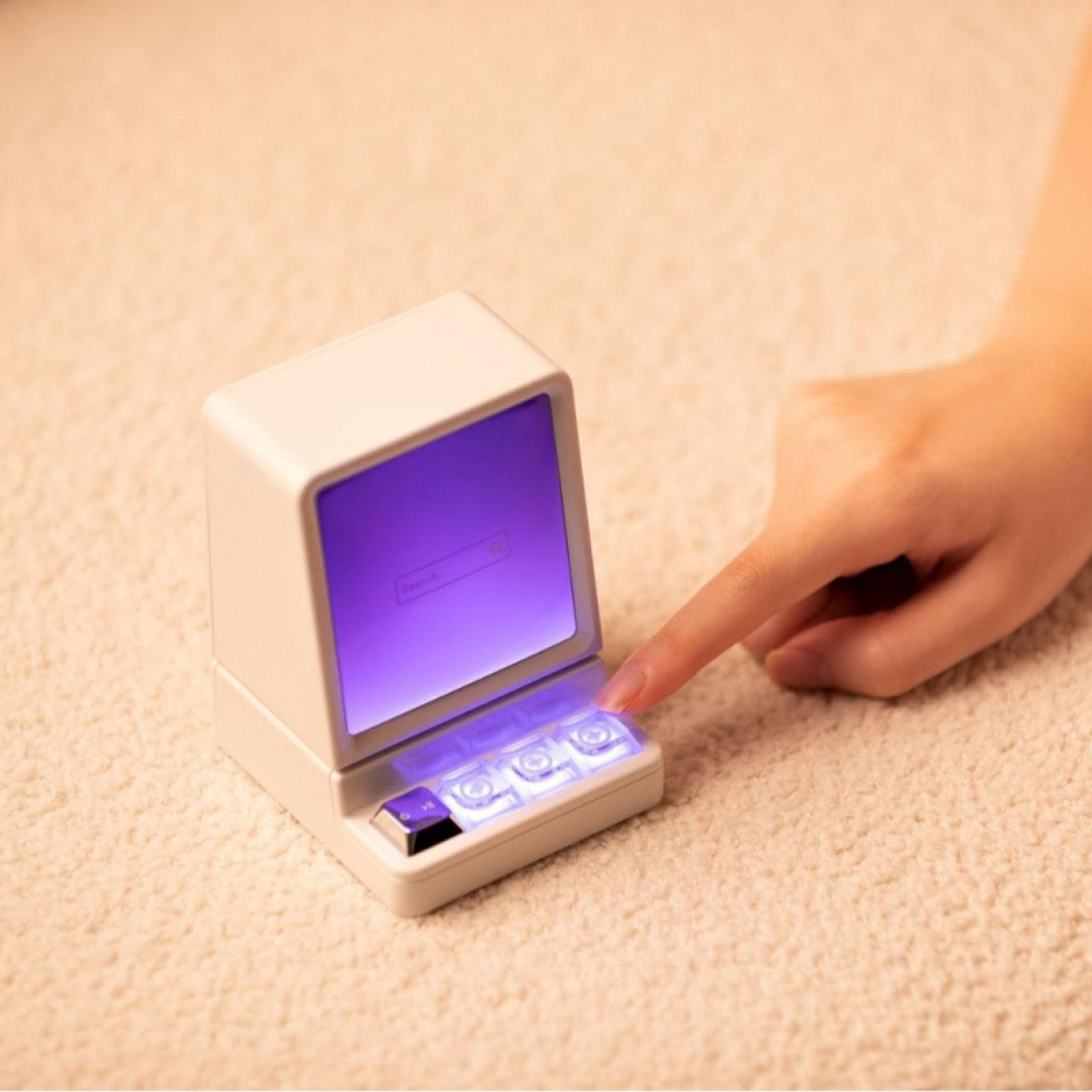 Ein Miniatur-Retro-Computer mit lila Bildschirm. Ein Finger berührt die beleuchteten Tasten. Beiger Teppich im Hintergrund.