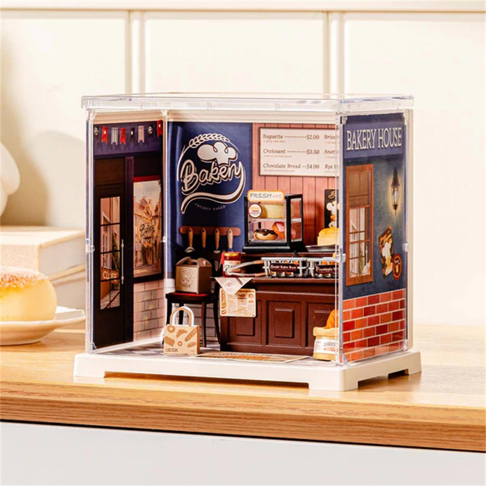 Miniatur-Bäckerei-Diorama mit Theke, Backwaren und Schild. Es befindet sich in einer transparenten Vitrine auf einer Holzoberfläche.