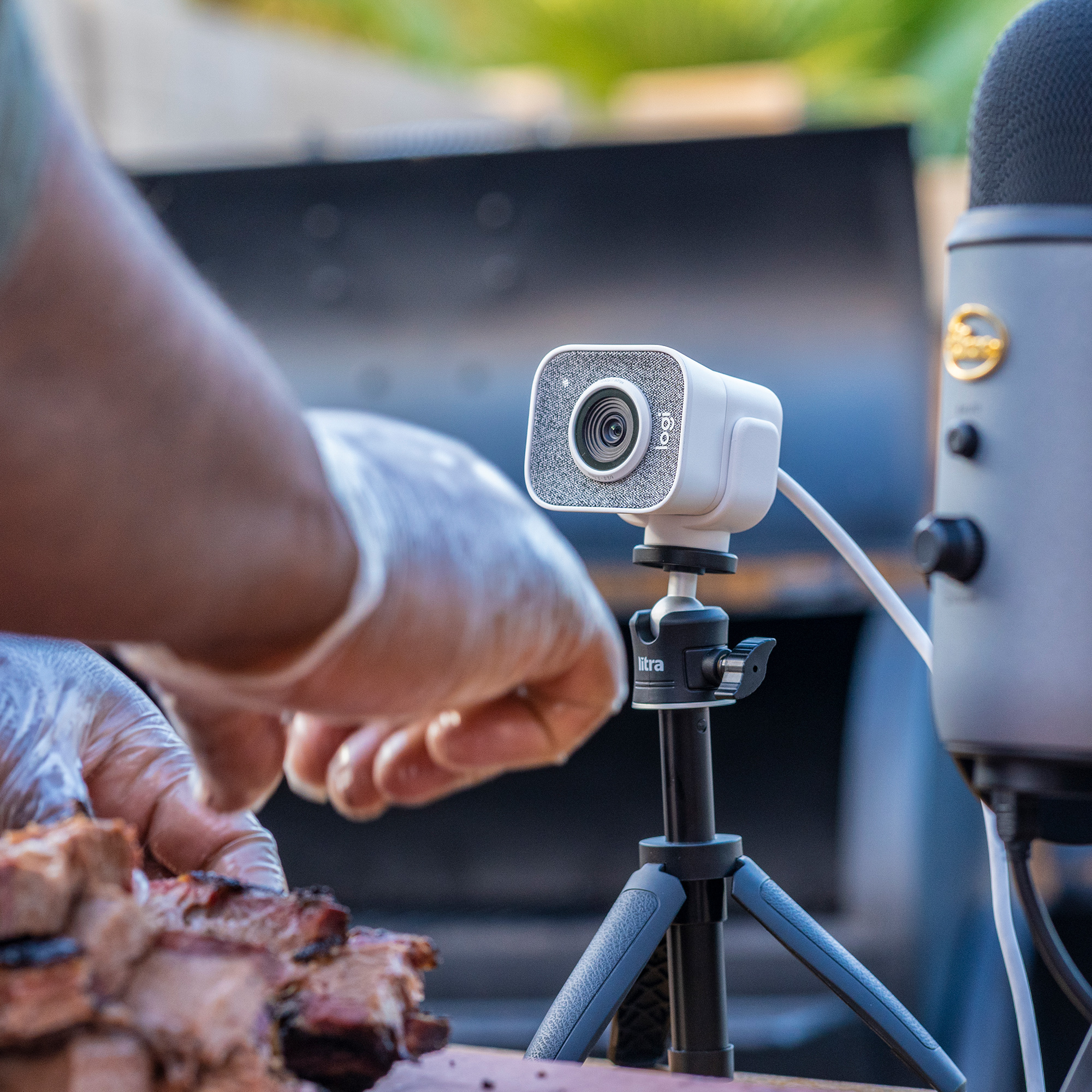 Kamera internetowa na statywie jest ustawiona w pobliżu mikrofonu i jedzenia. Widoczne są dłonie w rękawicach, przygotowujące jedzenie.
