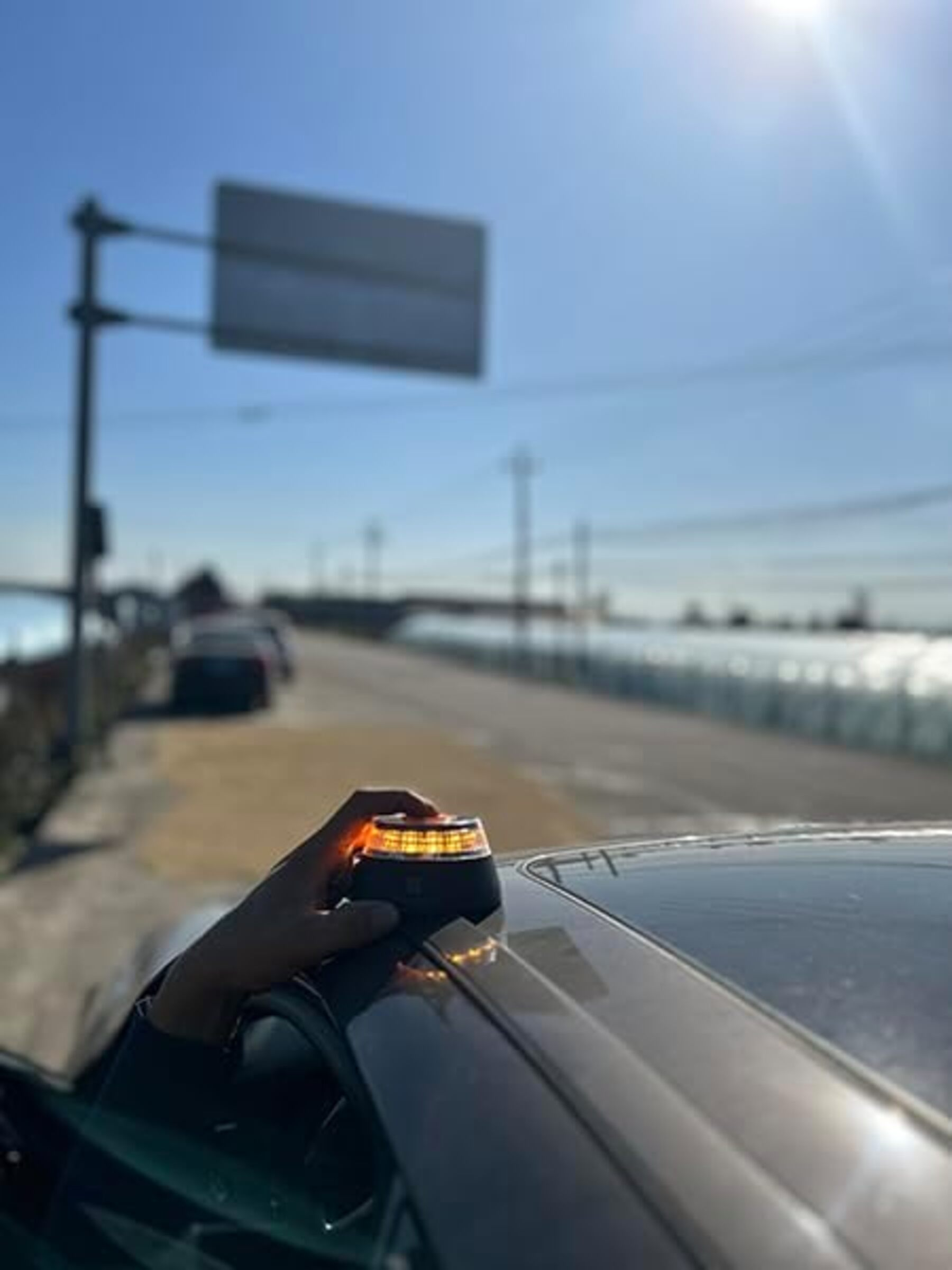 Una persona sostiene una luz negra en un coche. Un camino y un puente en el fondo bajo un cielo soleado.