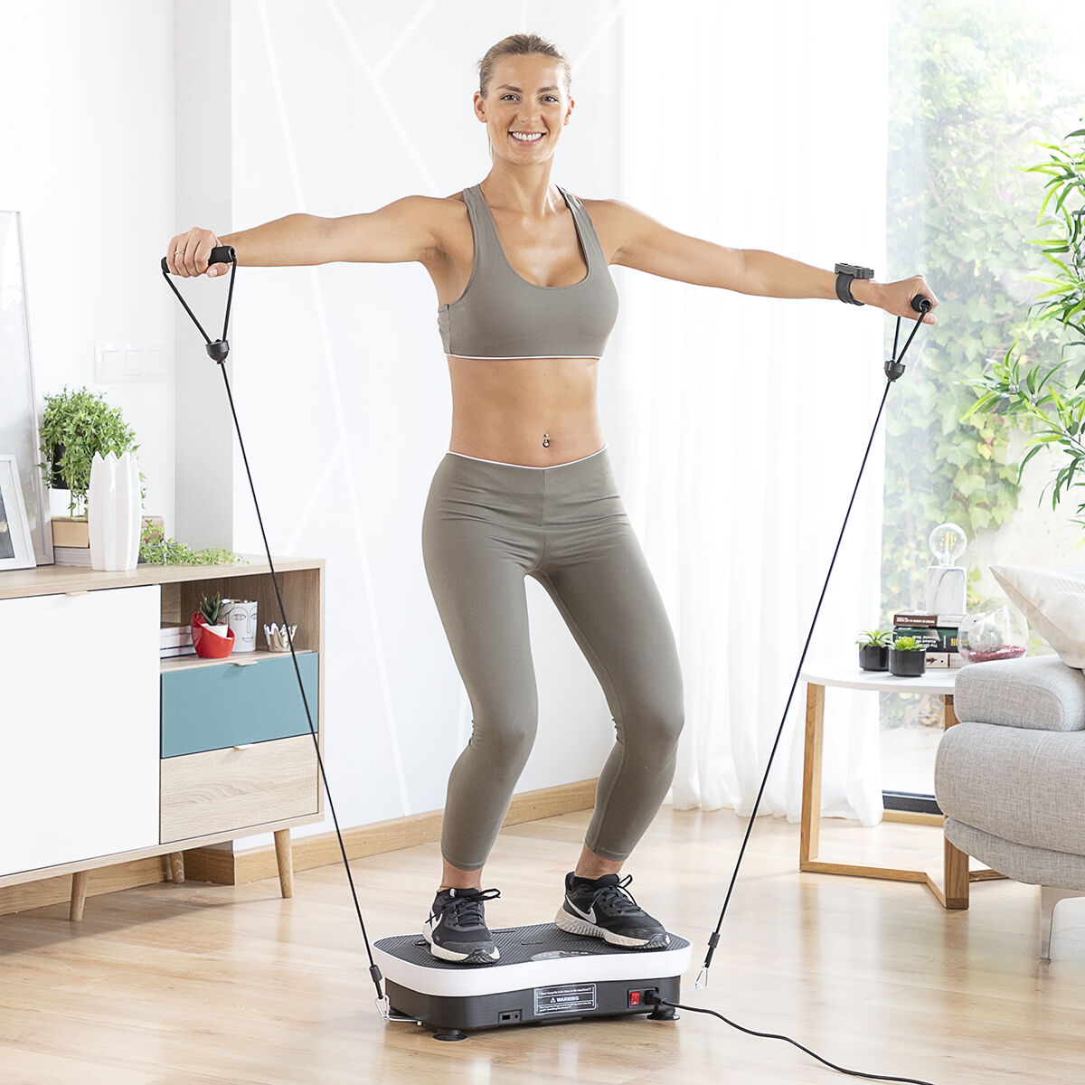 Femme sur une plateforme vibrante s'exerce avec des bandes. Elle porte des vêtements de sport. Arrière-plan : meubles, plantes et un canapé.