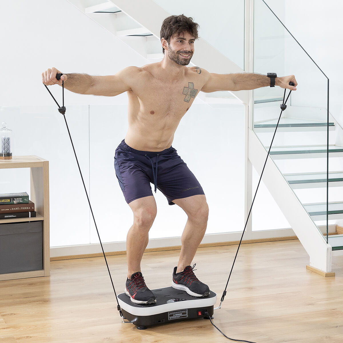 Un homme s'exerce sur une plateforme vibrante, tenant des bandes de résistance. Il porte un short et des chaussures. Escalier et étagère en arrière-plan.