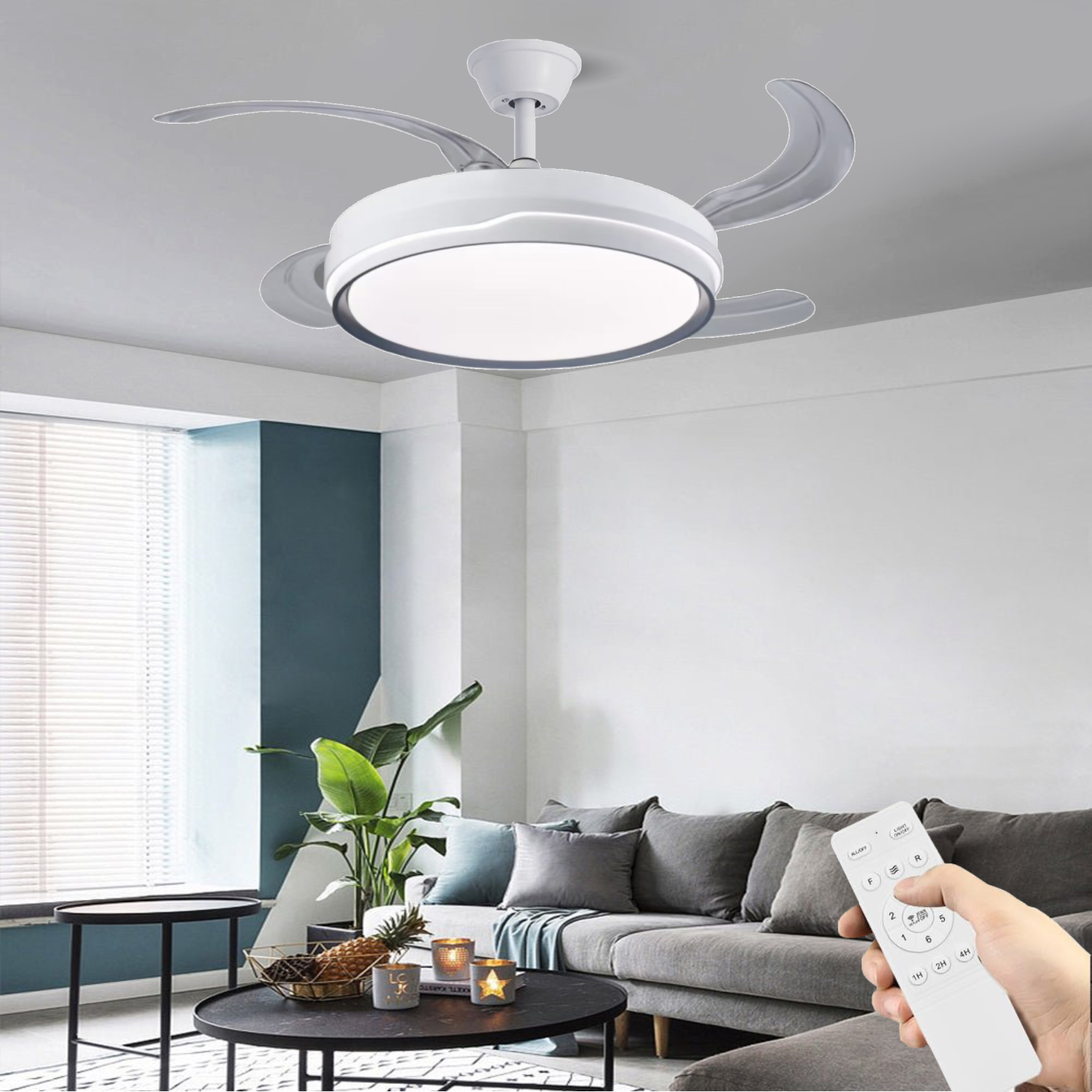 Ventilador de techo blanco con luz en una sala de estar. Una mano sostiene un mando a distancia. Sofá gris, mesas redondas negras y plantas.