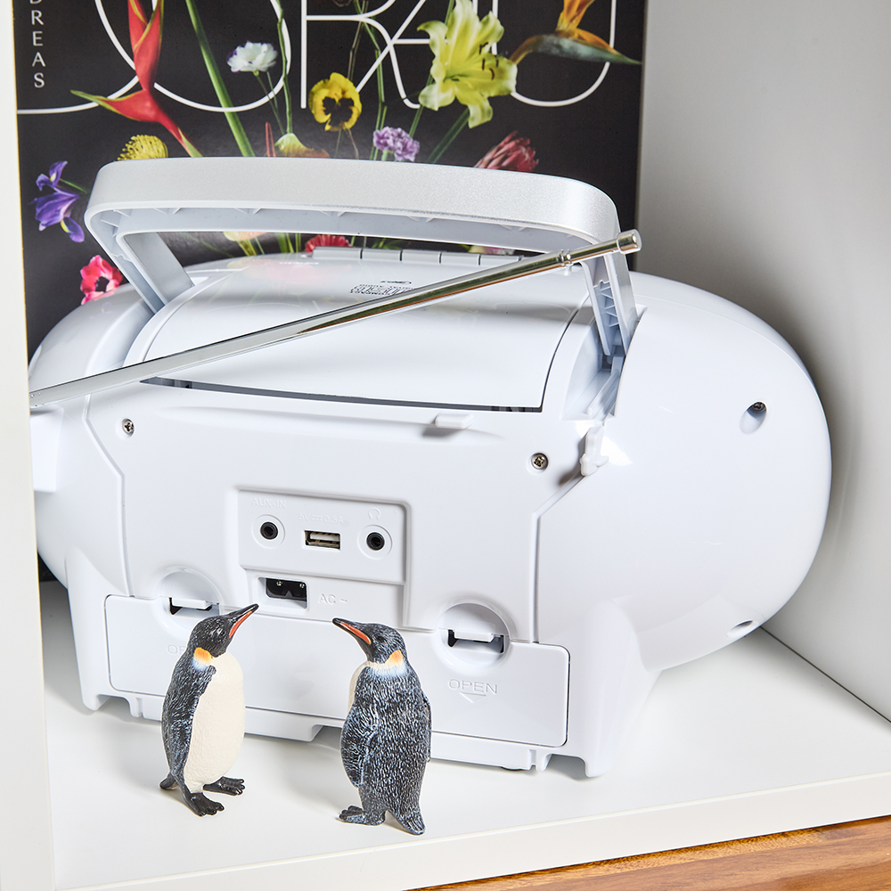 Ein weißer Ghettoblaster mit silberner Antenne und Griff, zwei Pinguinfiguren stehen davor, eine Zeitschrift mit Blumen im Hintergrund.