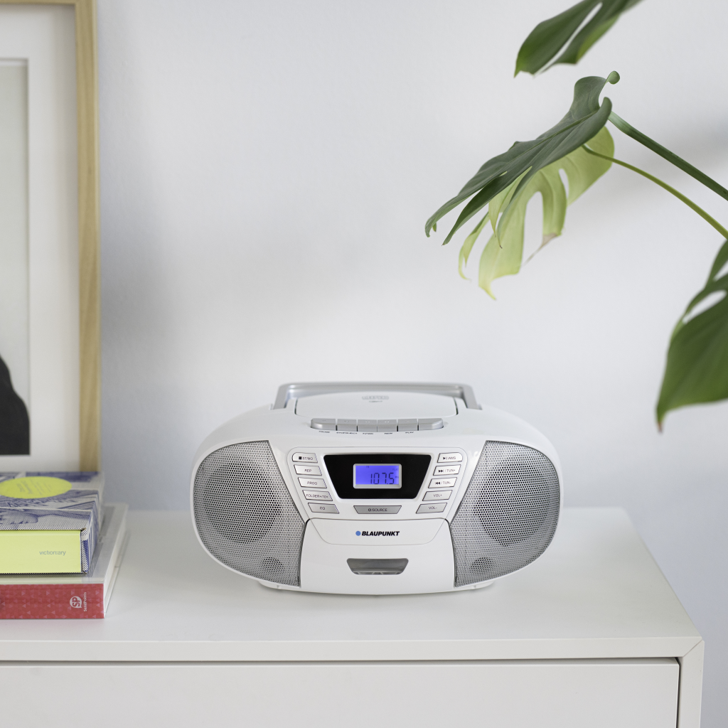 Weißer Ghettoblaster mit Digitalanzeige auf einem weißen Tisch. Grüne Pflanze im Hintergrund. Bücher links, gerahmte Kunst hinten.