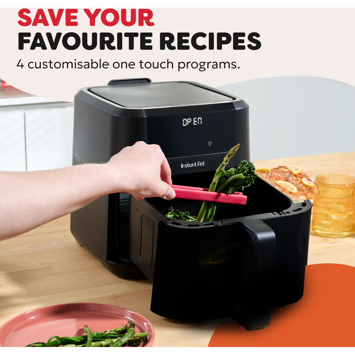 Een hand plaatst asperges in een zwarte airfryer. Een roze bord met asperges en een glas water staan op tafel.