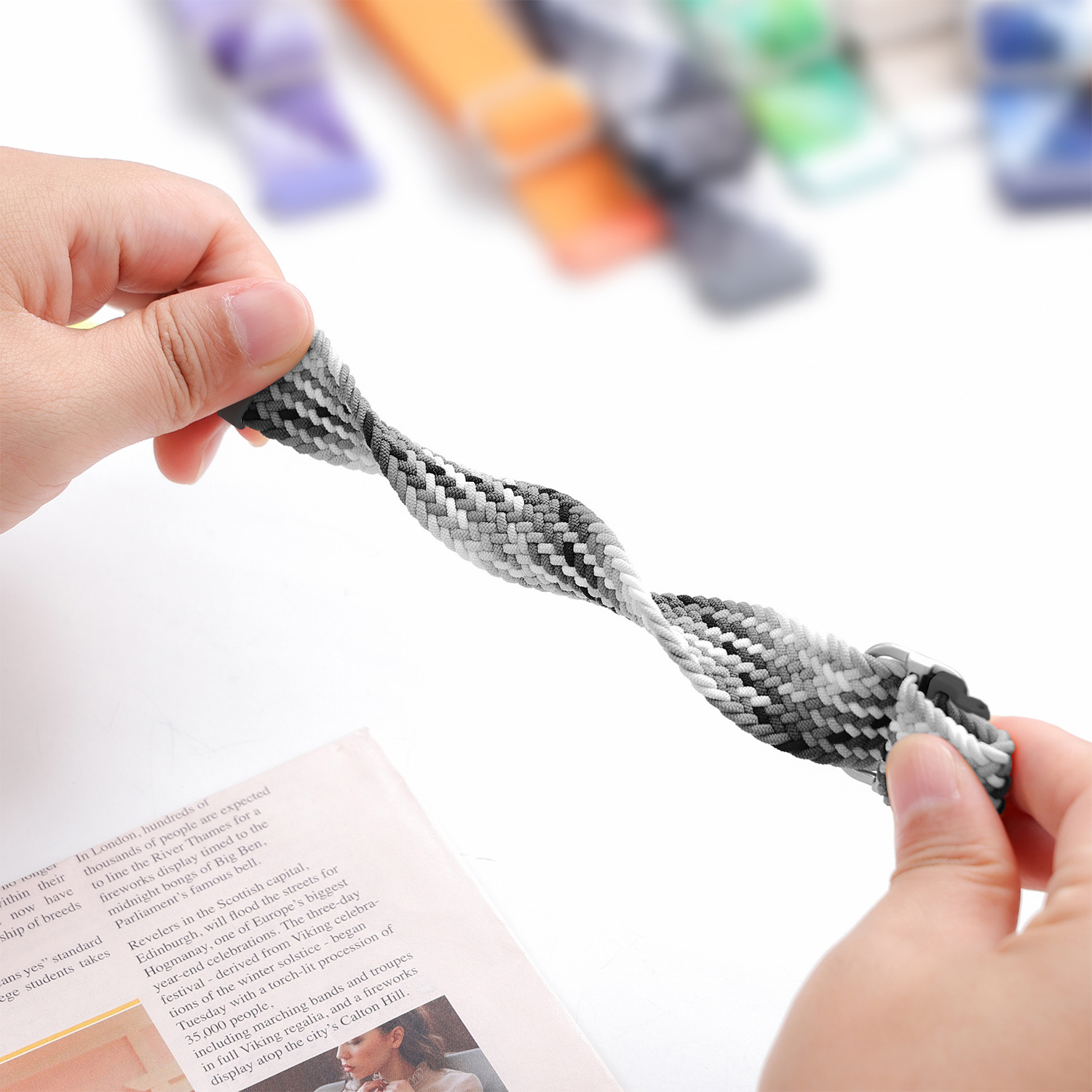 Dłonie trzymające plecioną bransoletkę w kolorze szarym i białym. Tło jest rozmyte, pokazujące inne bransoletki i gazetę.