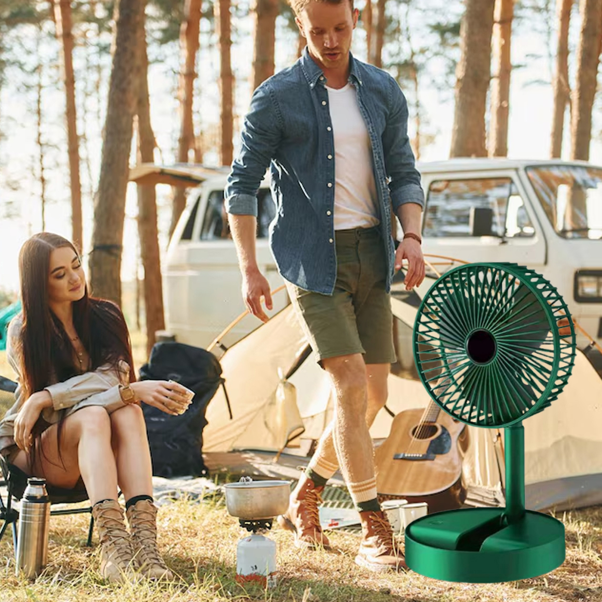 Ein Paar beim Camping im Wald. Im Vordergrund steht ein grüner Ventilator. Eine Frau sitzt und hält eine Tasse. Ein Mann geht. Zelt und Van sind im Hintergrund.