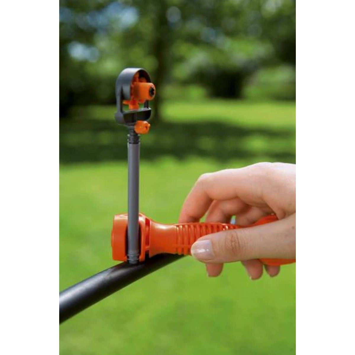 Ręka trzyma pomarańczowy zraszacz przymocowany do czarnej rury. W tle widoczna jest zielona trawa i drzewa.