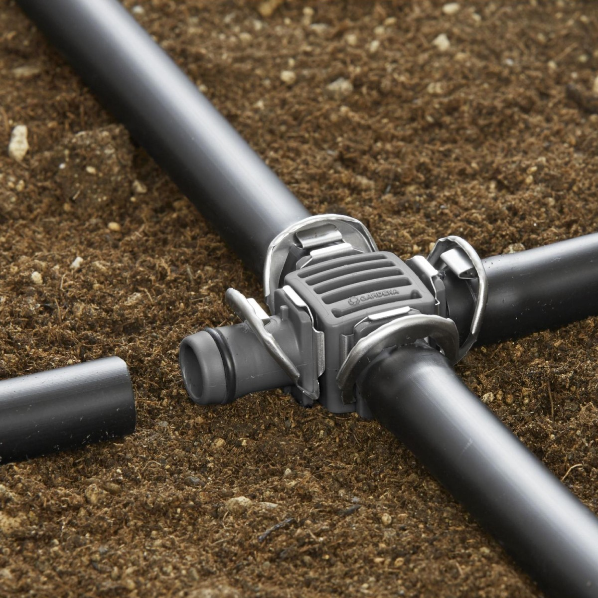 Un conector Gardena gris une tubos negros sobre tierra marrón. El conector tiene forma cuadrada con clips metálicos.