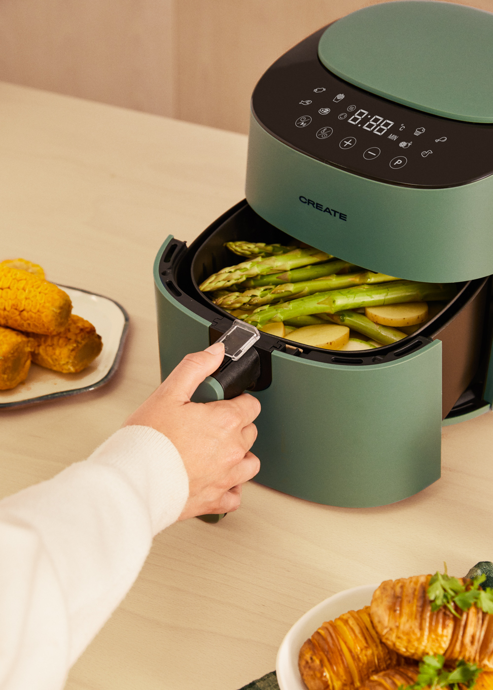 Una freidora de aire verde con espárragos y patatas dentro. Una mano tira del cajón. Maíz y patatas en platos.