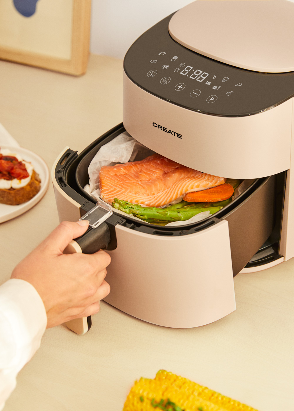 Ein Airfryer mit Lachs und Gemüse, eine Hand zieht den Korb heraus. Auf der linken Seite ein Teller mit Essen.