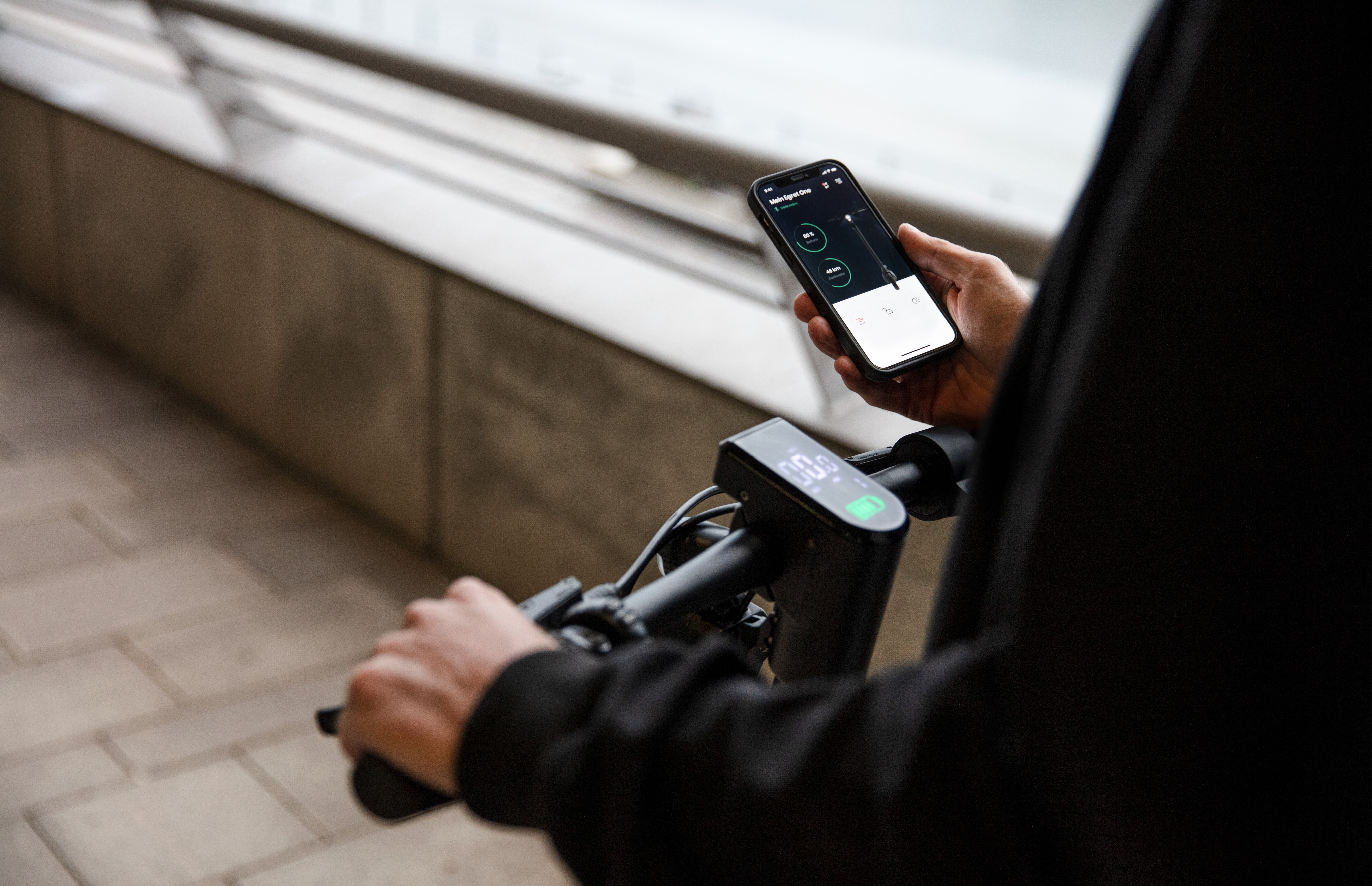 Eine Person hält ein Telefon mit Rollerinformationen. Der Lenker des Rollers ist im Fokus. Der Hintergrund ist unscharf.