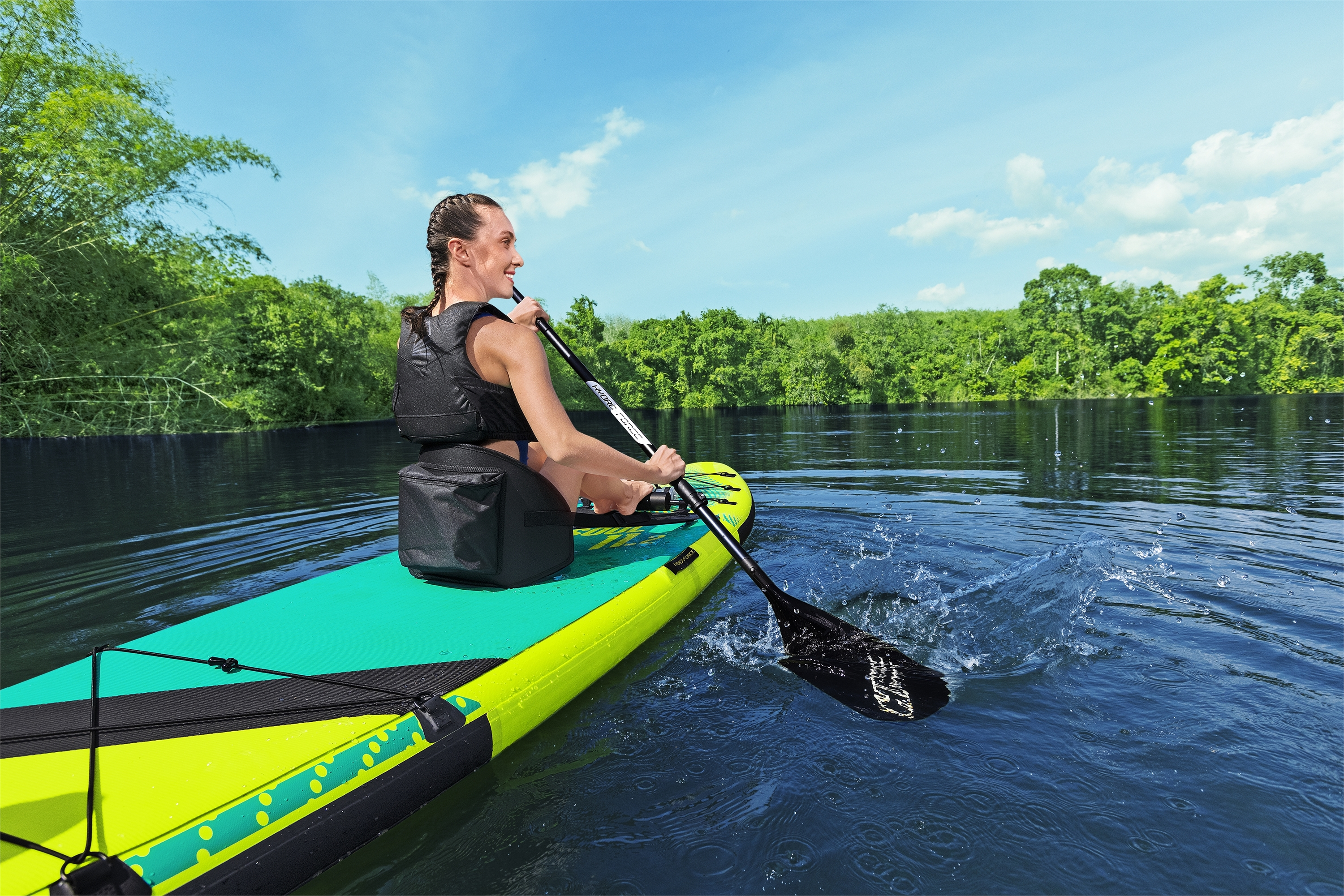 Vrouw peddelt op een paddleboard op een meer. Ze draagt een zwemvest. Groene bomen en blauwe lucht op de achtergrond.