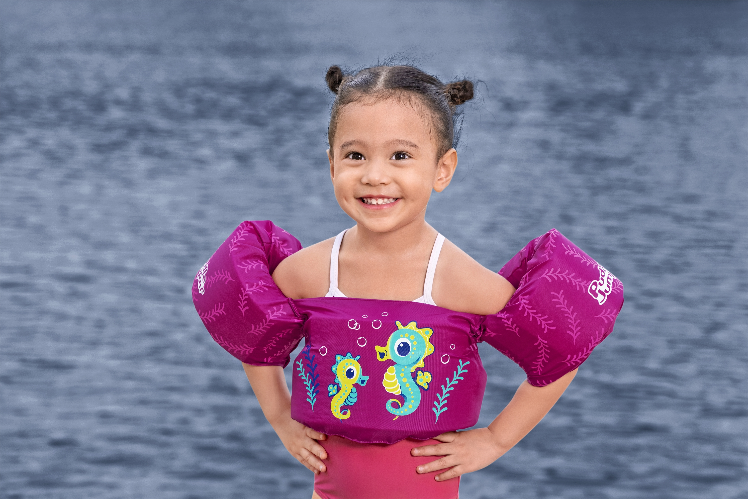Una bambina con un costume da bagno rosa e braccioli sorride, posando davanti a uno sfondo d'acqua.