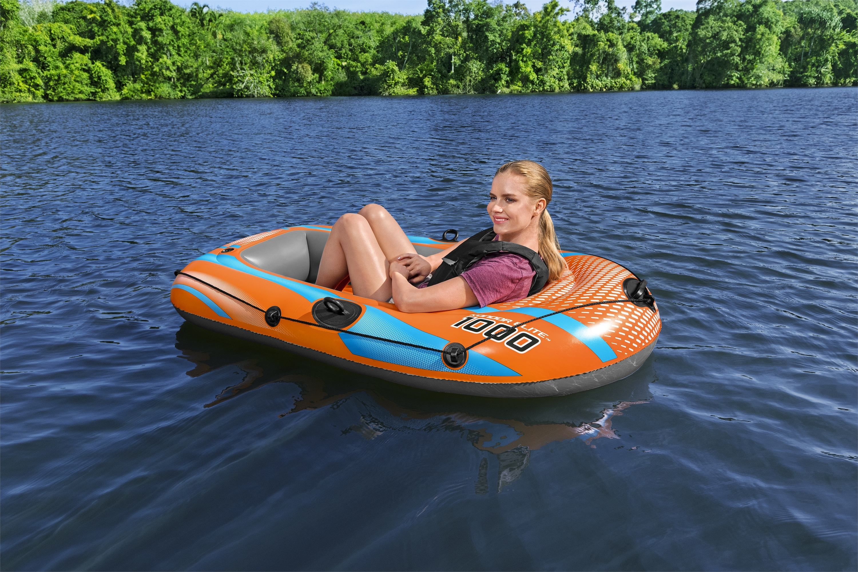 Eine Frau sitzt in einem orangefarbenen und blauen Schlauchboot auf einem See. Bäume säumen das ferne Ufer.