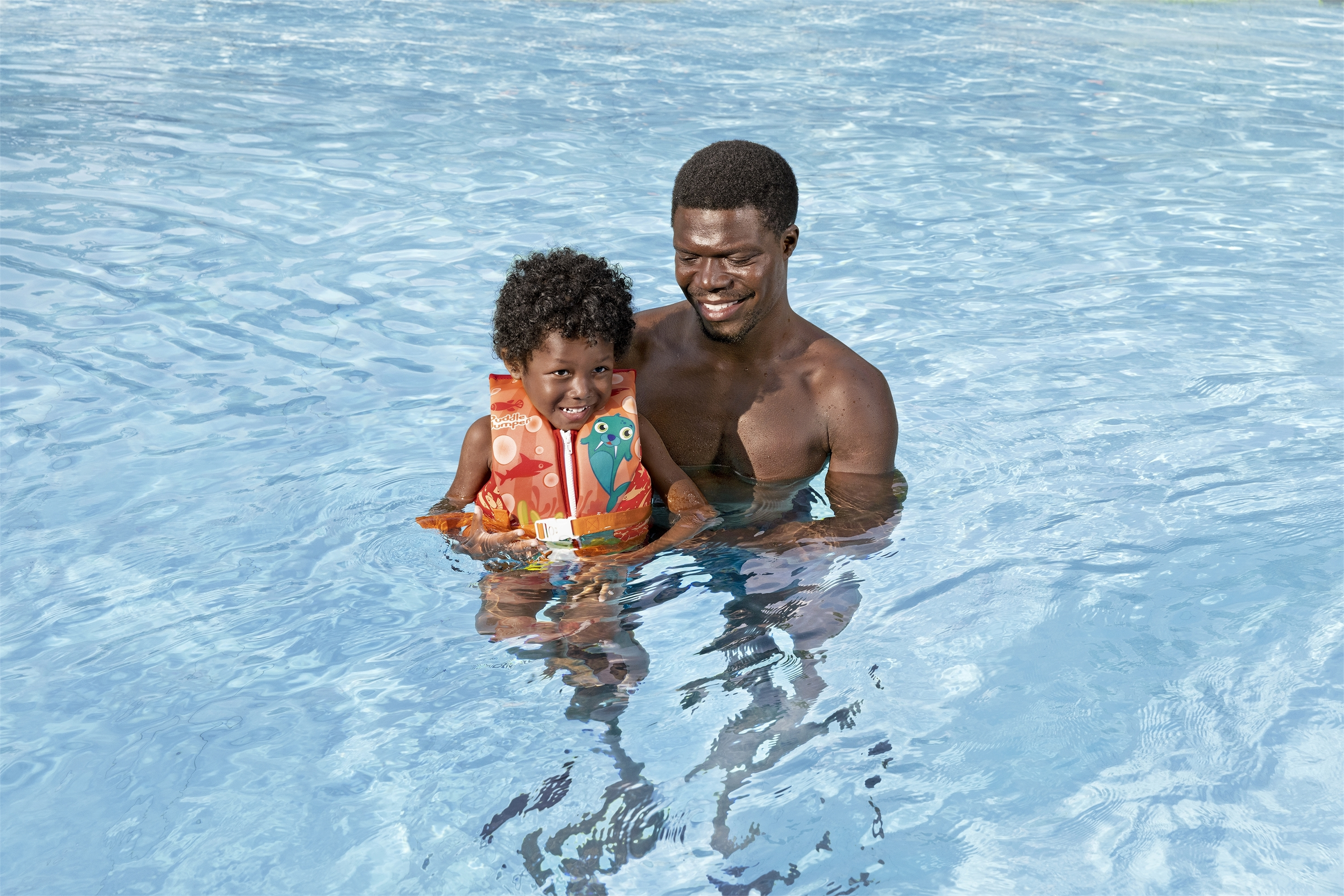 Un uomo e un bambino in piscina. Il bambino indossa un giubbotto di salvataggio. Entrambi sorridono. L'acqua è blu.