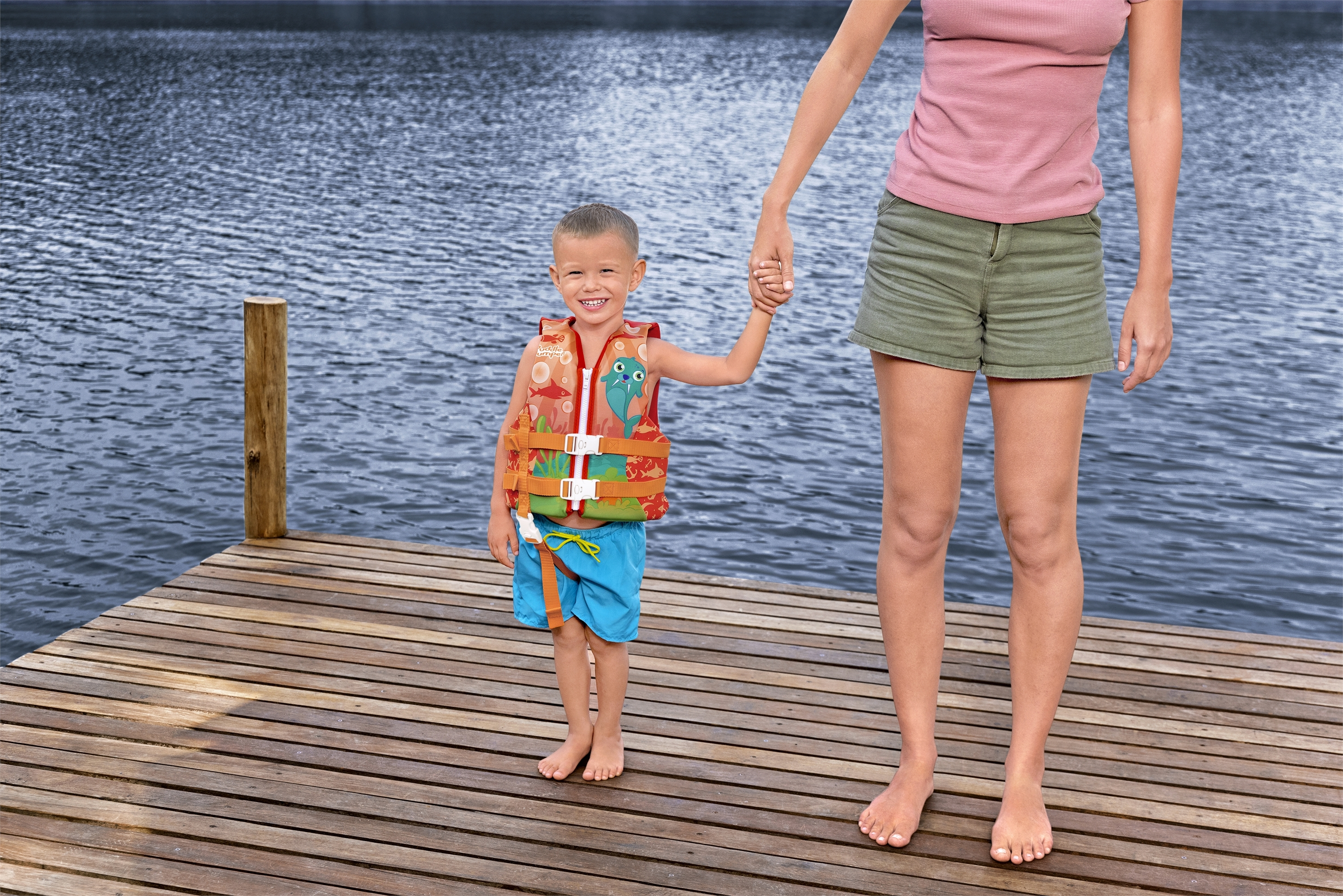 Un bambino con un giubbotto di salvataggio tiene la mano di una donna su un pontile di legno. Lo sfondo è un lago.