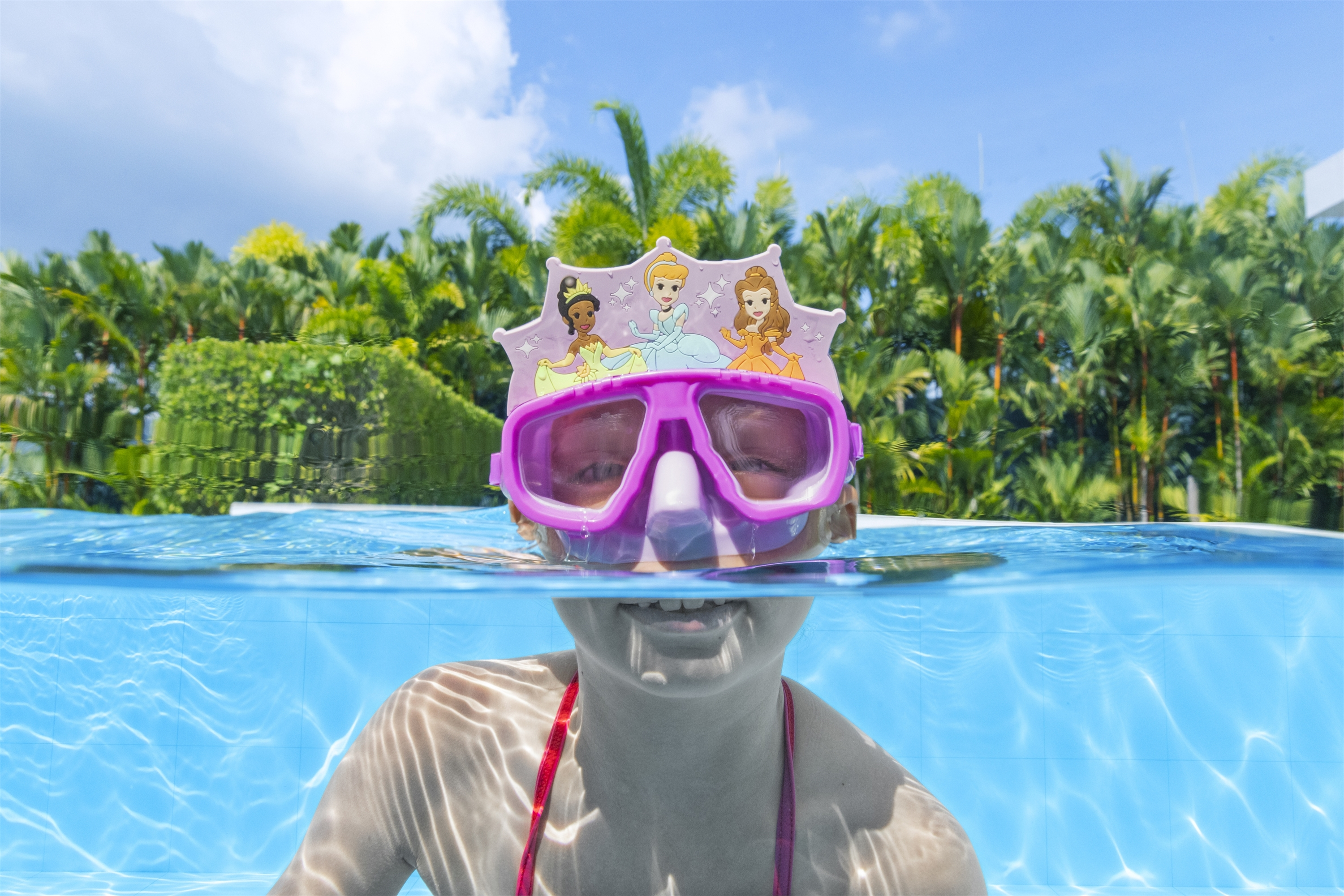Una persona con una maschera subacquea a tema principesse è in piscina. Sullo sfondo ci sono lussureggianti alberi verdi e cielo blu.