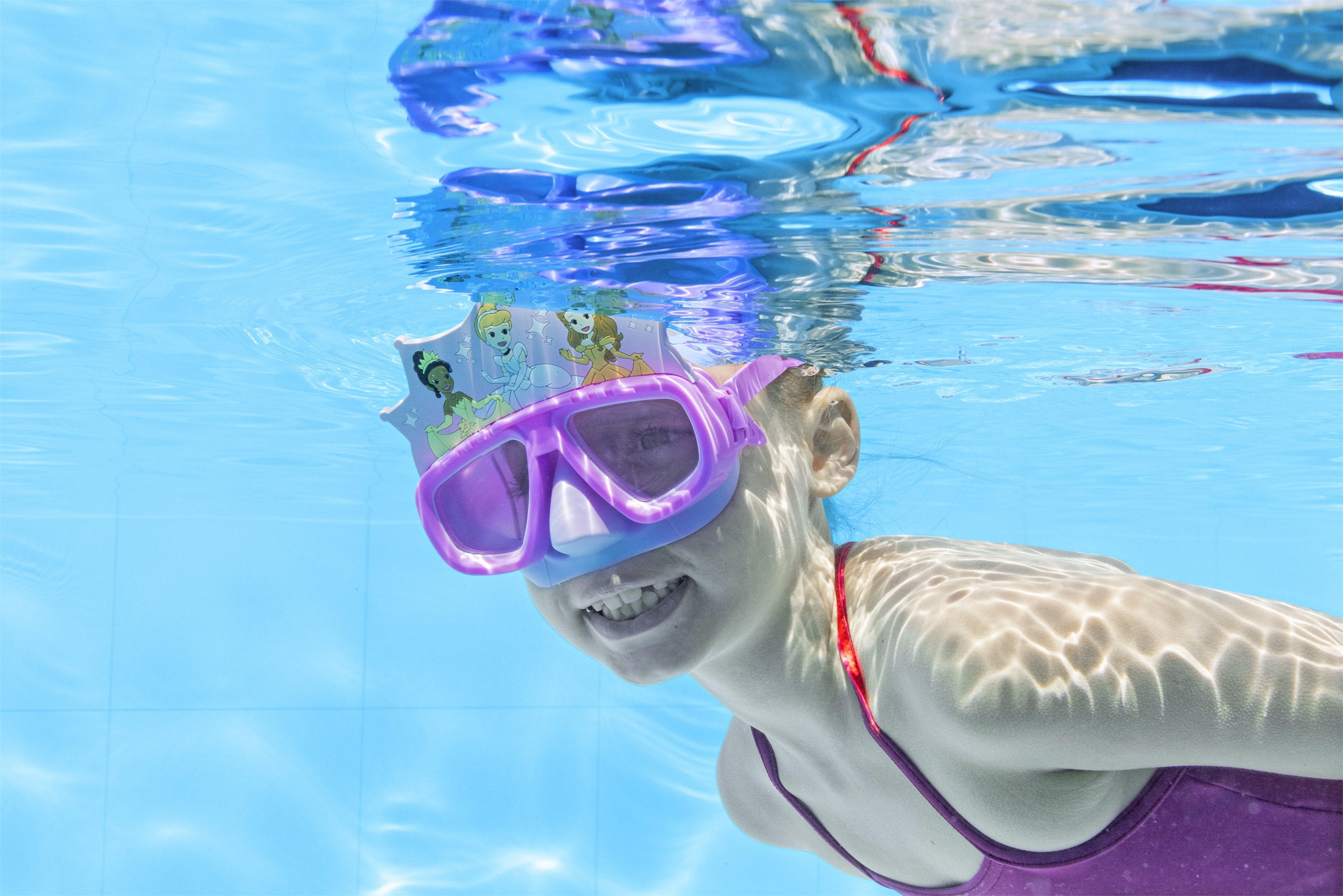 Una persona sott'acqua con occhiali viola e una cuffia da nuoto a tema principesse. Lo sfondo è acqua blu.