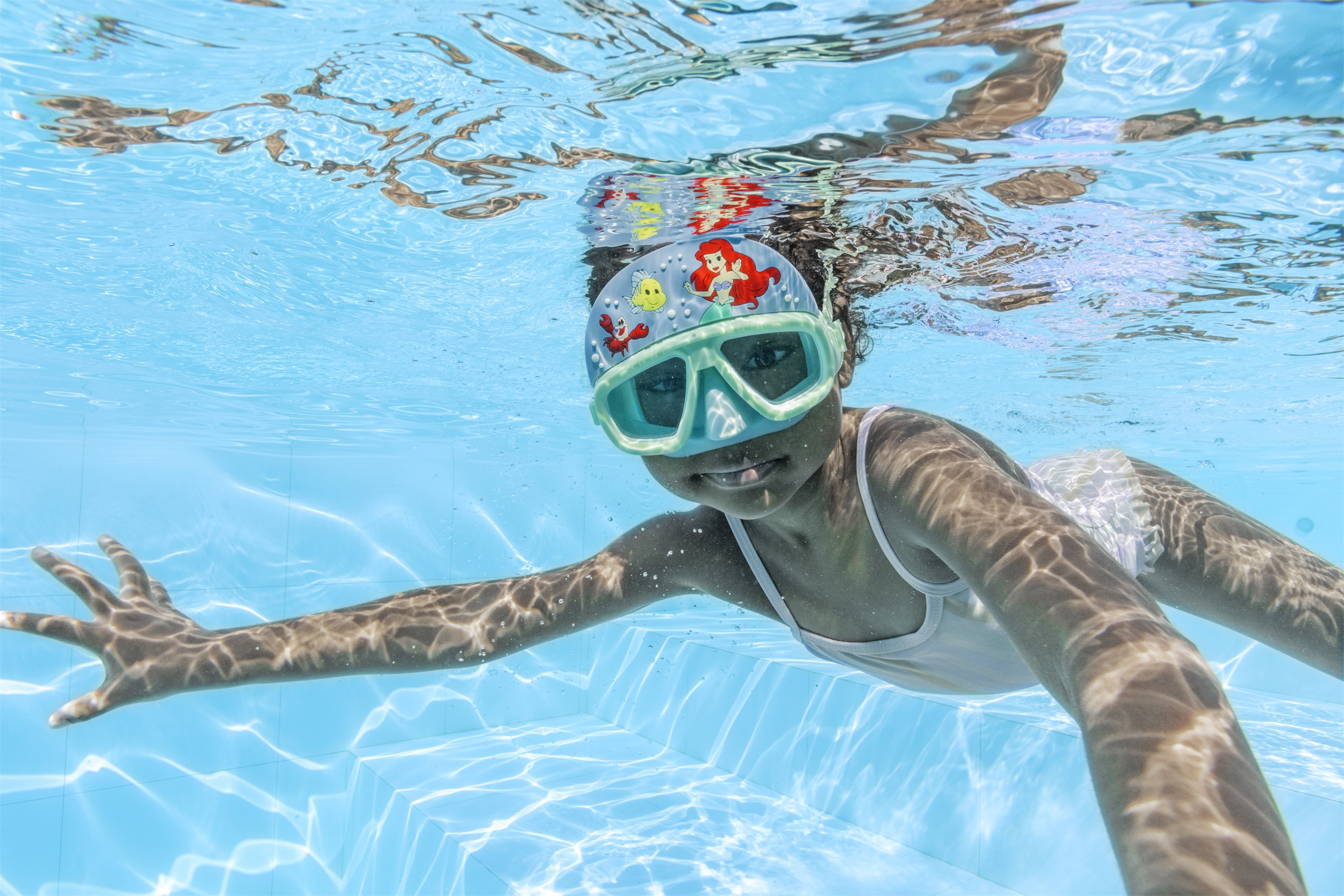 Un bambino sott'acqua in piscina, con occhialini e cuffia. Il bambino allunga un braccio.