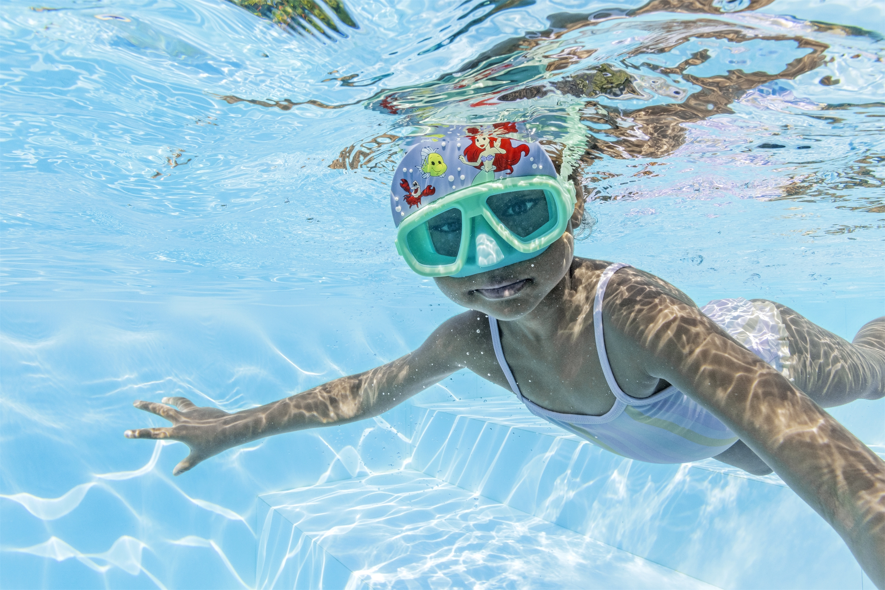 Un bambino nuota sott'acqua in una piscina, con occhialini e cuffia. La luce solare si riflette nell'acqua.