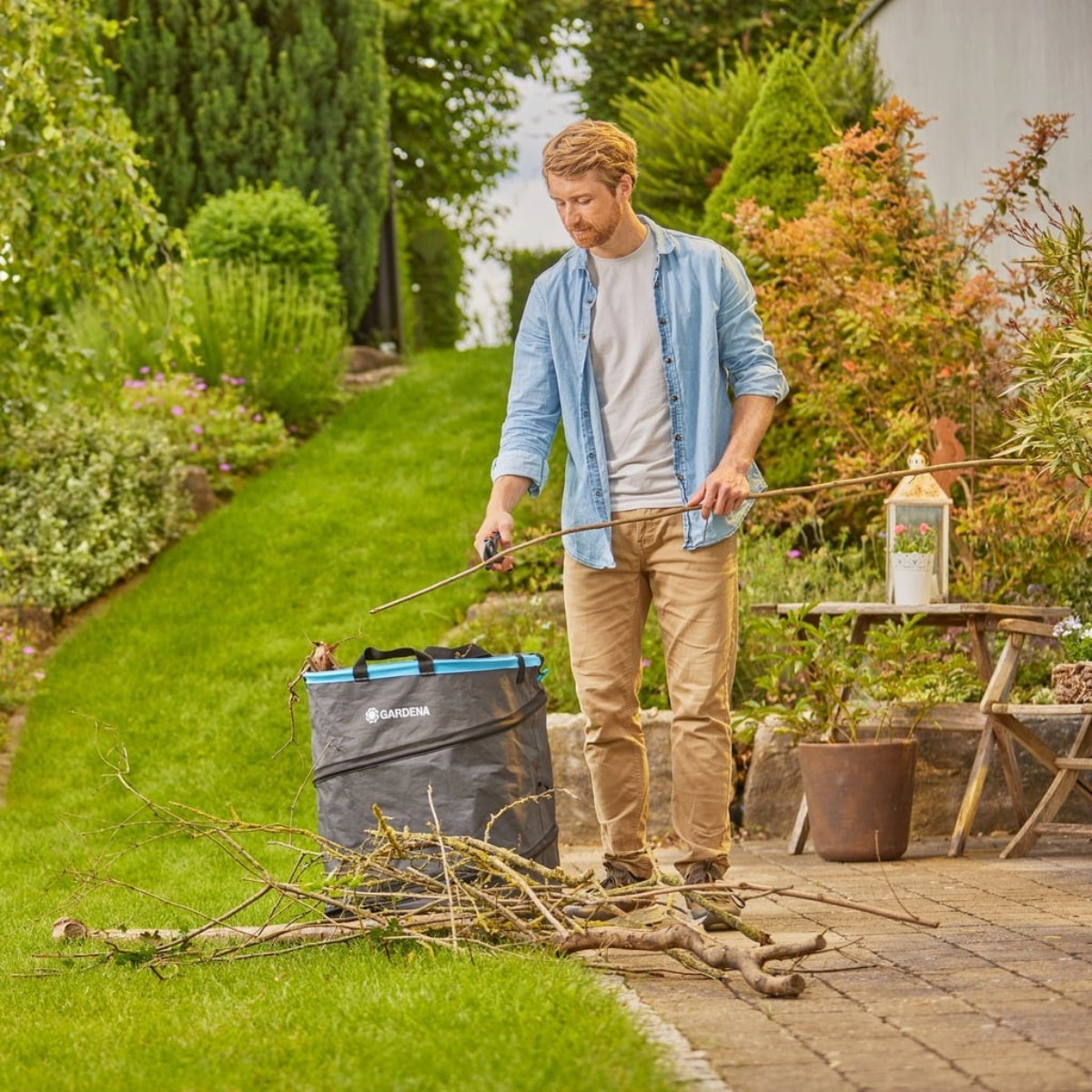 Mężczyzna w ogrodzie trzyma gałęzie, umieszczając je w szarej torbie. Zielona trawa i rośliny w tle.