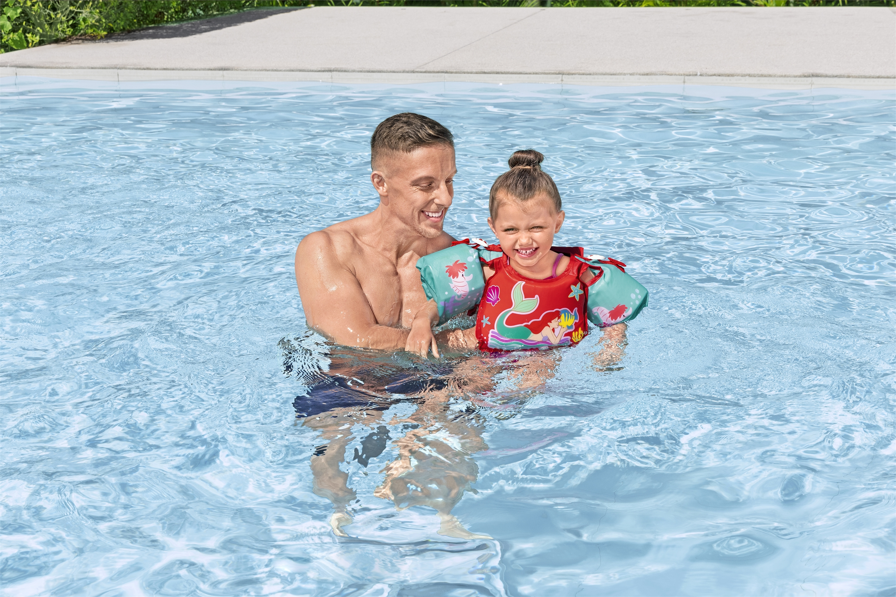 Un uomo e una bambina sono in piscina. La bambina indossa un giubbotto di salvataggio. Entrambi sorridono.