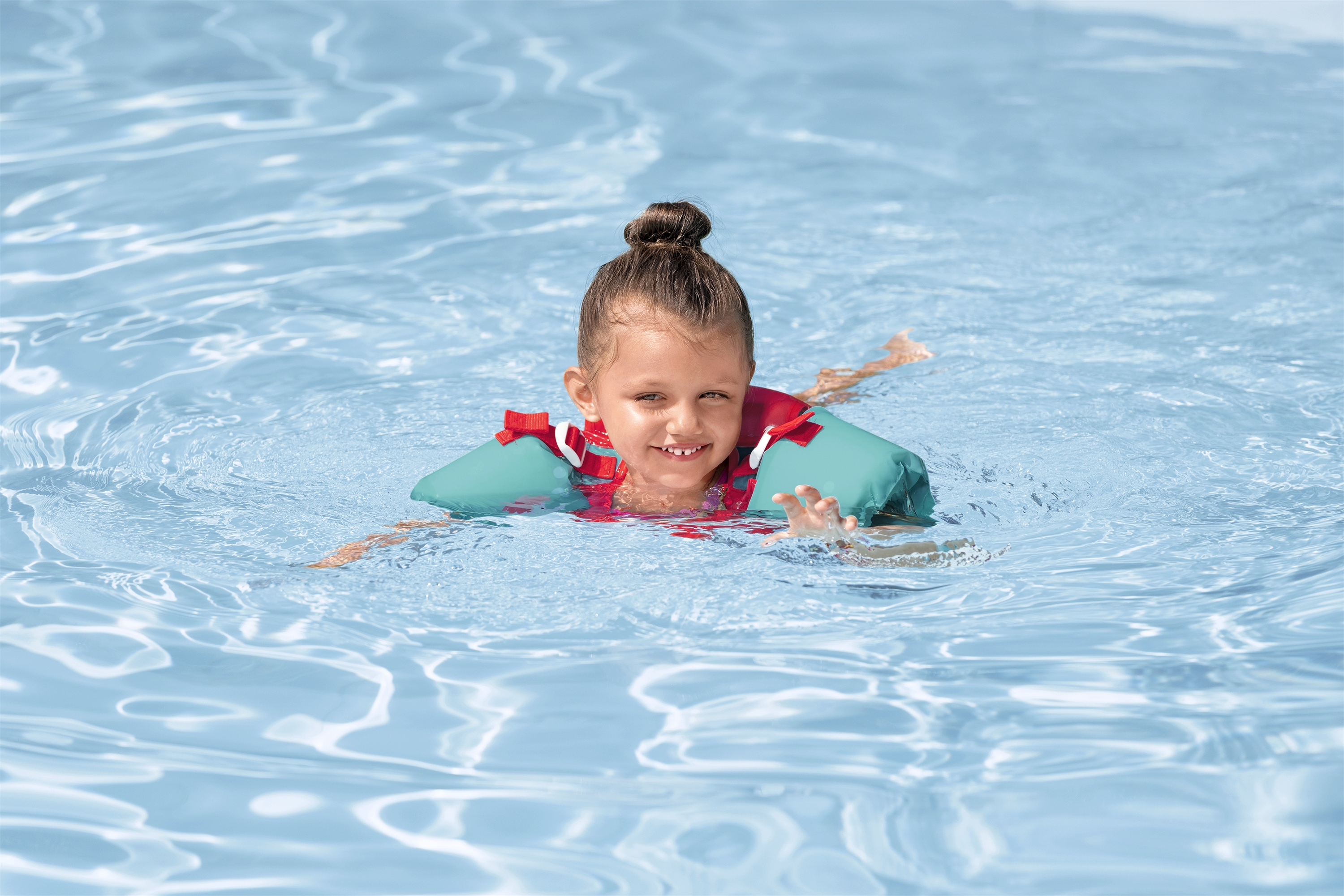 Una ragazzina con uno chignon nuota in una piscina indossando un giubbotto di salvataggio verde acqua e rosso, sorridendo alla telecamera.