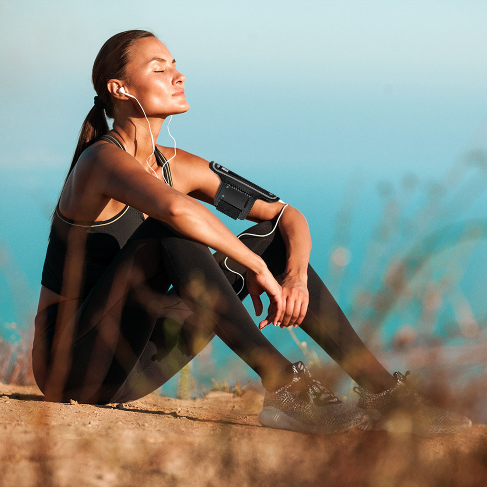 Femme en tenue de sport assise dehors, yeux fermés, brassard avec téléphone. Fond : ciel bleu et nature floue.