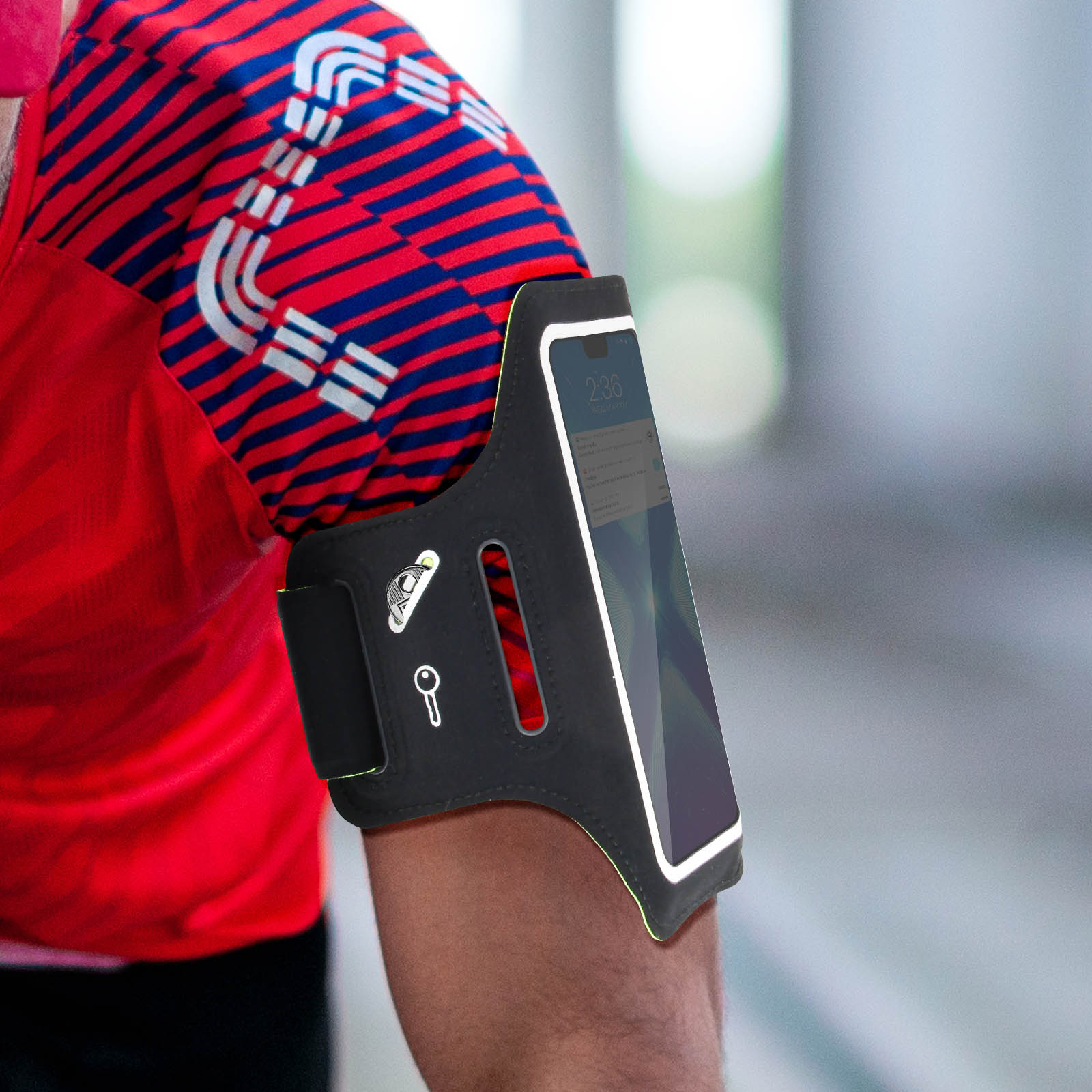 Un bras avec un téléphone dans un brassard noir. Chemise rouge avec design bleu et blanc. Le téléphone affiche 2:36.