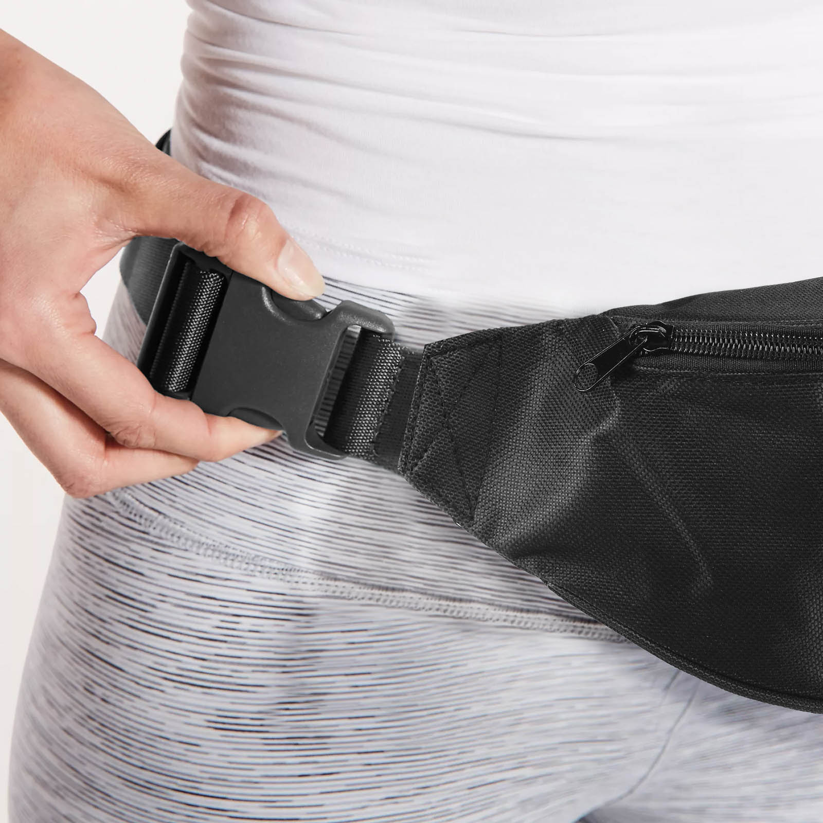 Die Hand einer Person befestigt eine schwarze Bauchtasche. Die Person trägt ein weißes Oberteil und graue Leggings.