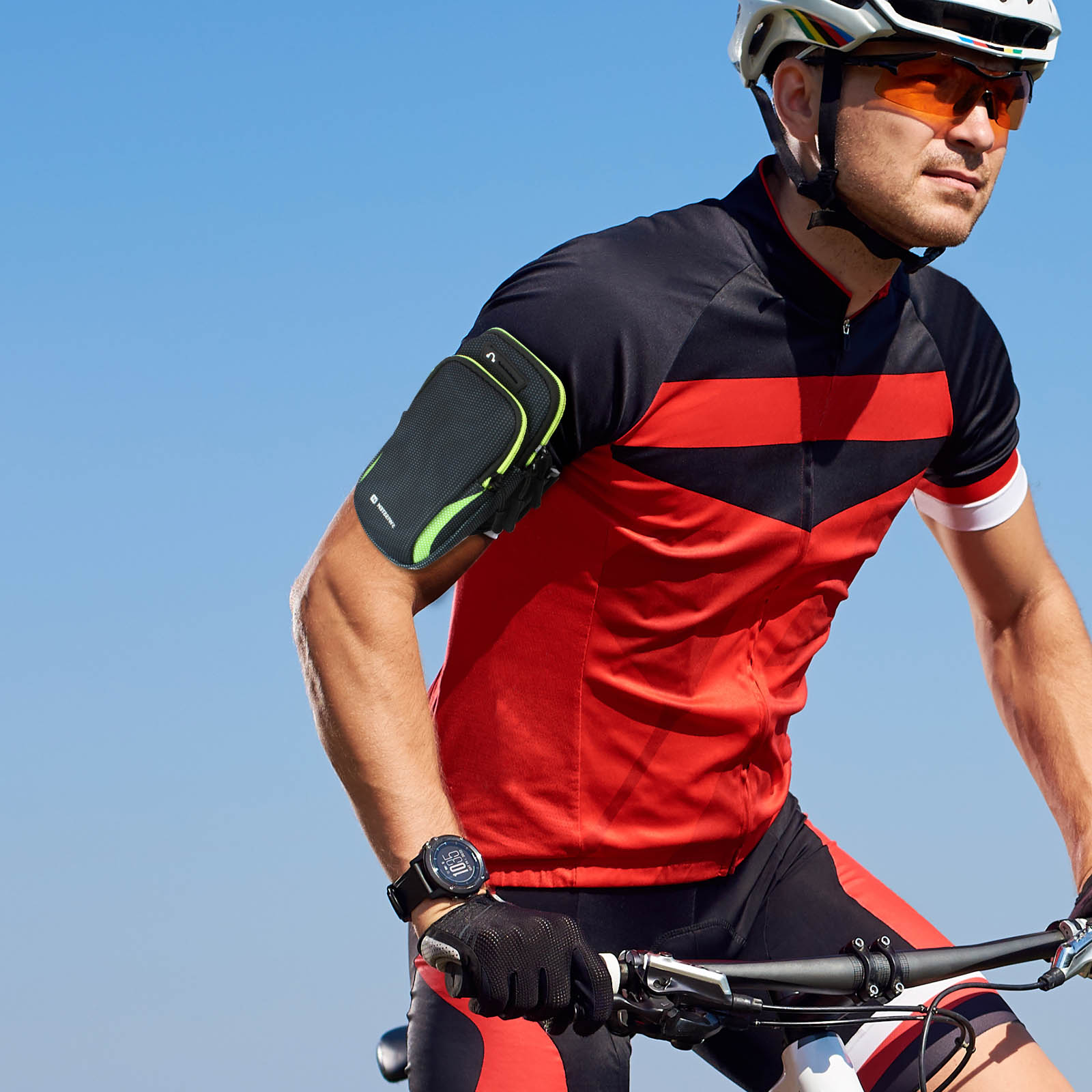 Cycliste à vélo portant un casque, des lunettes de soleil et un maillot rouge/noir. Un brassard maintient un téléphone. Ciel bleu clair en arrière-plan.
